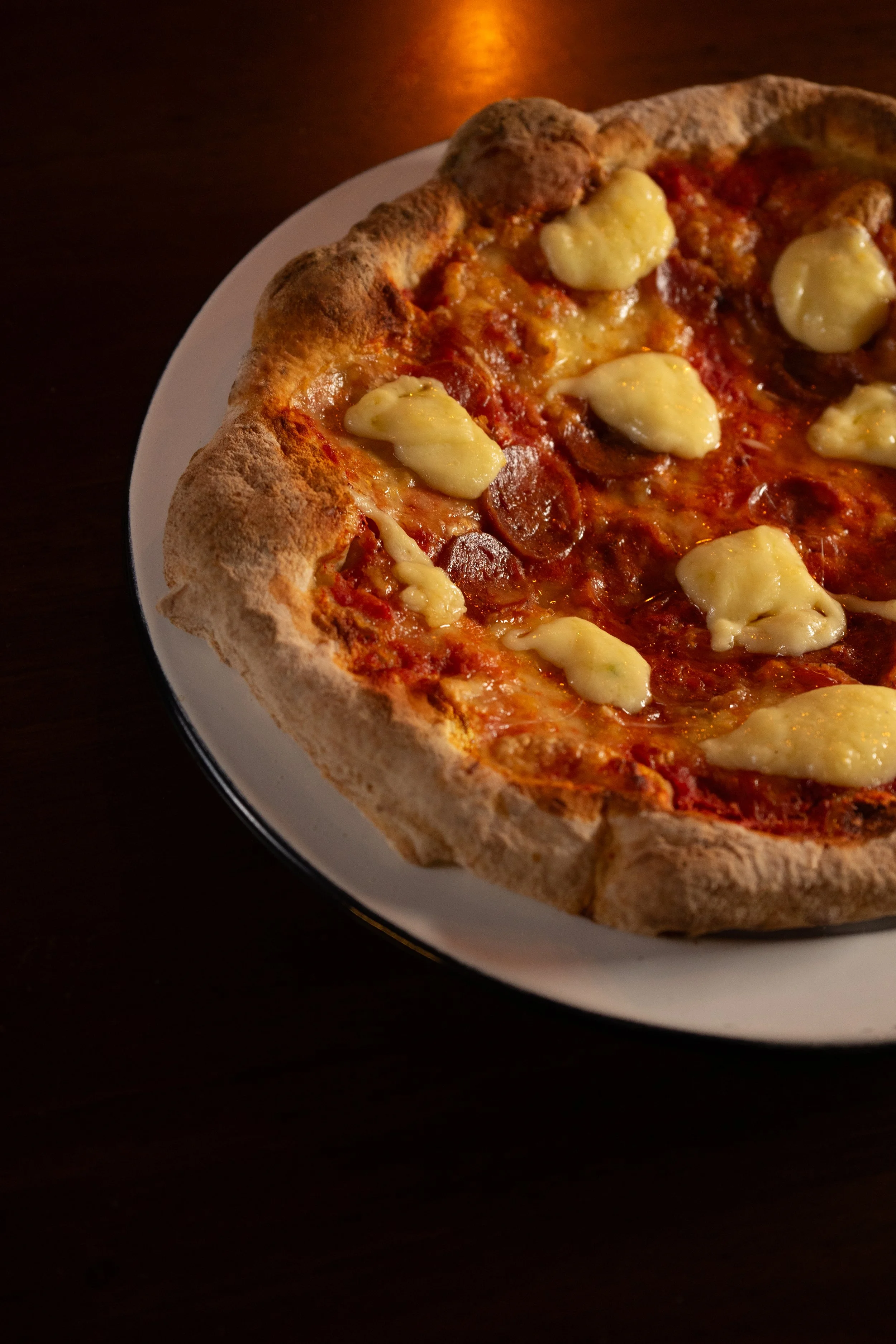 Pizza con queso, pepperoni y masa gruesa en un plato blanco sobre fondo de madera oscura.