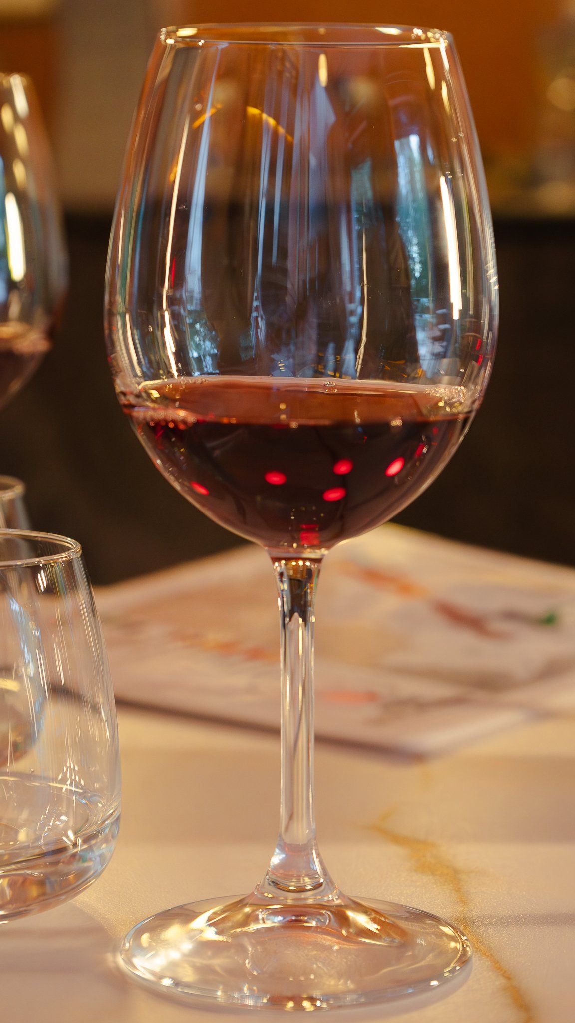 Una copa de vino tinto en un ambiente de restaurante, con reflejos de luces y otros vasos en el fondo.