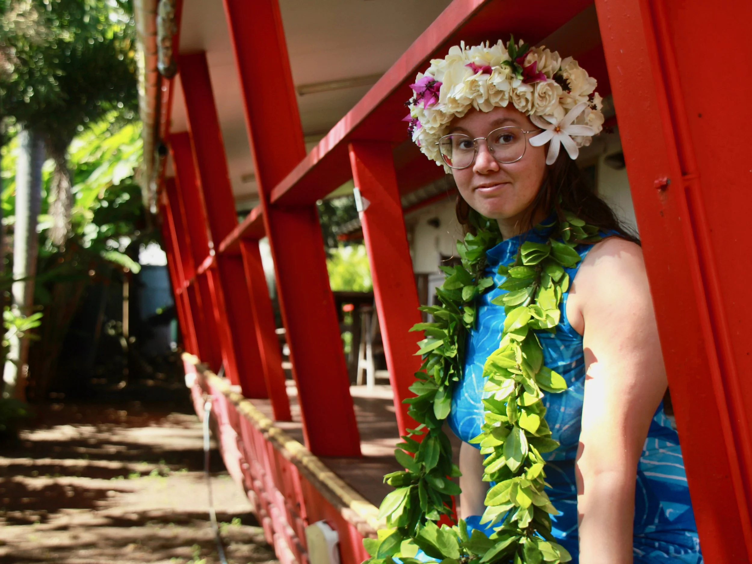 Young woman with glasses wearing a floral crown and green lei, standing inside a red structure with a lush outdoor setting in the background.