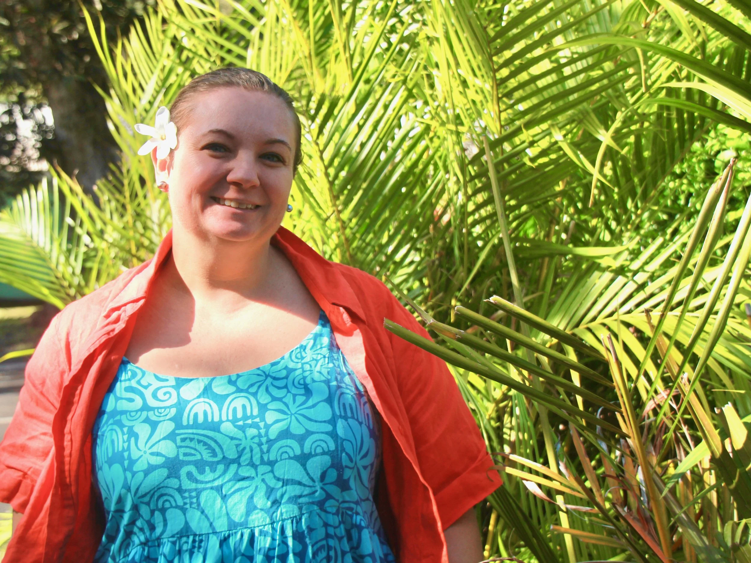A smiling woman with a flower in her hair, wearing a blue patterned dress and an orange shirt, standing outdoors amongst lush green tropical plants.