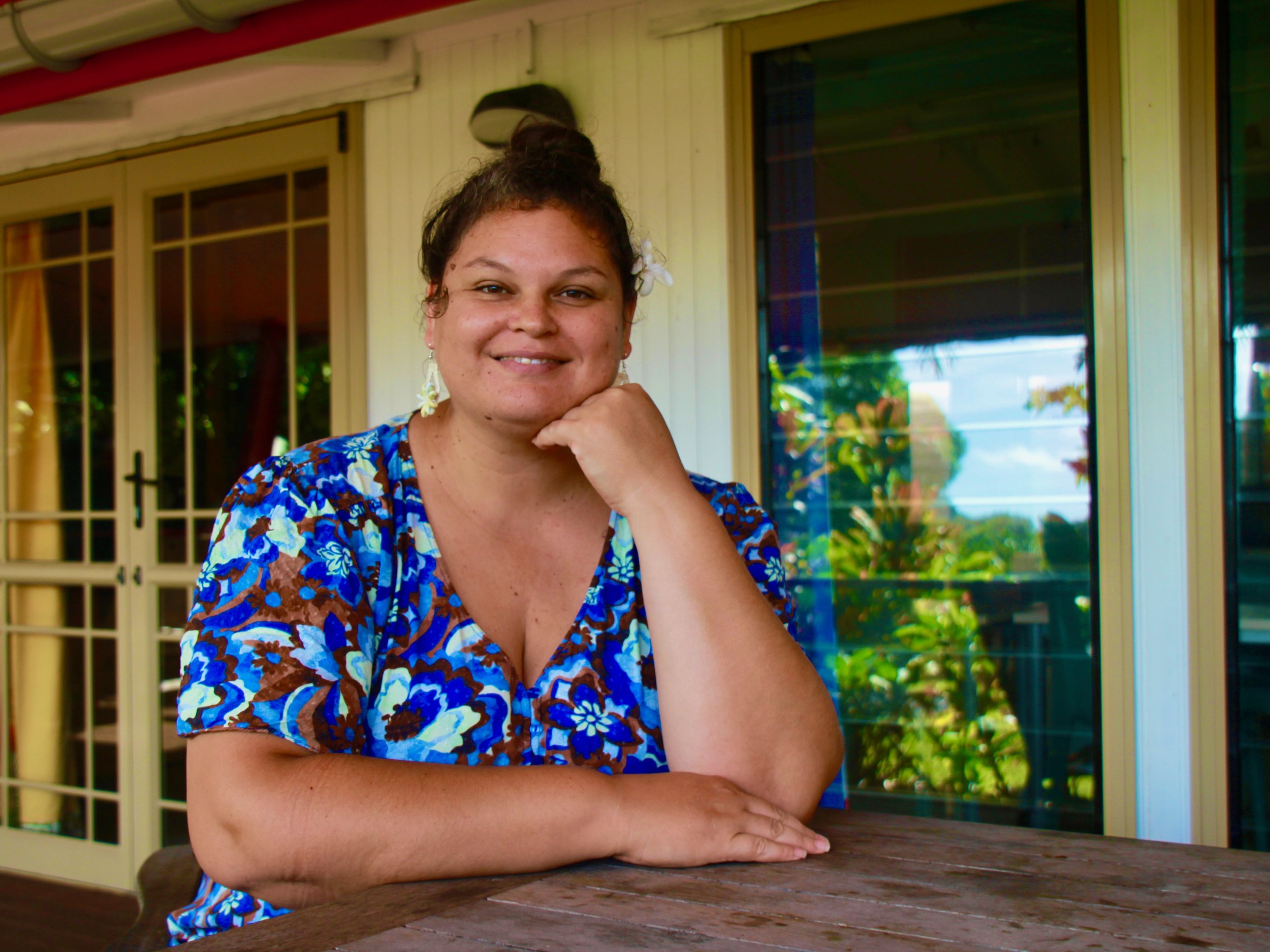 A woman with brown hair tied up, wearing a blue floral top, sitting at a table with her chin resting on her hand, smiling outdoors in front of a house with large glass windows and a sliding door, reflecting trees and sky.