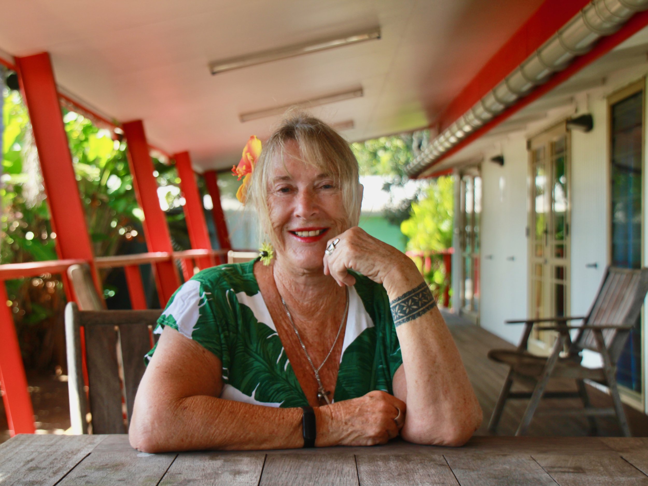 An elderly woman with blonde hair and a flower in her hair sitting at a table on a porch, smiling and resting her chin on her hand, with lush greenery in the background.
