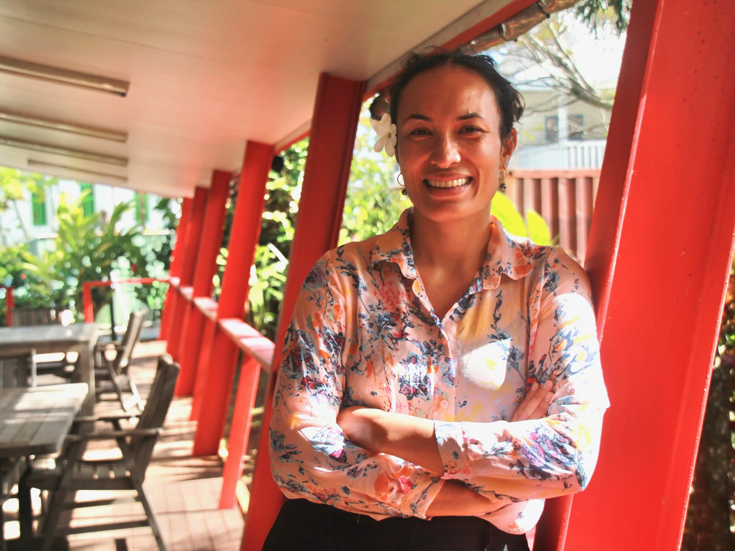 Woman with a flower in her hair smiling with arms crossed, standing on a patio with red columns and outdoor furniture, greenery in the background.