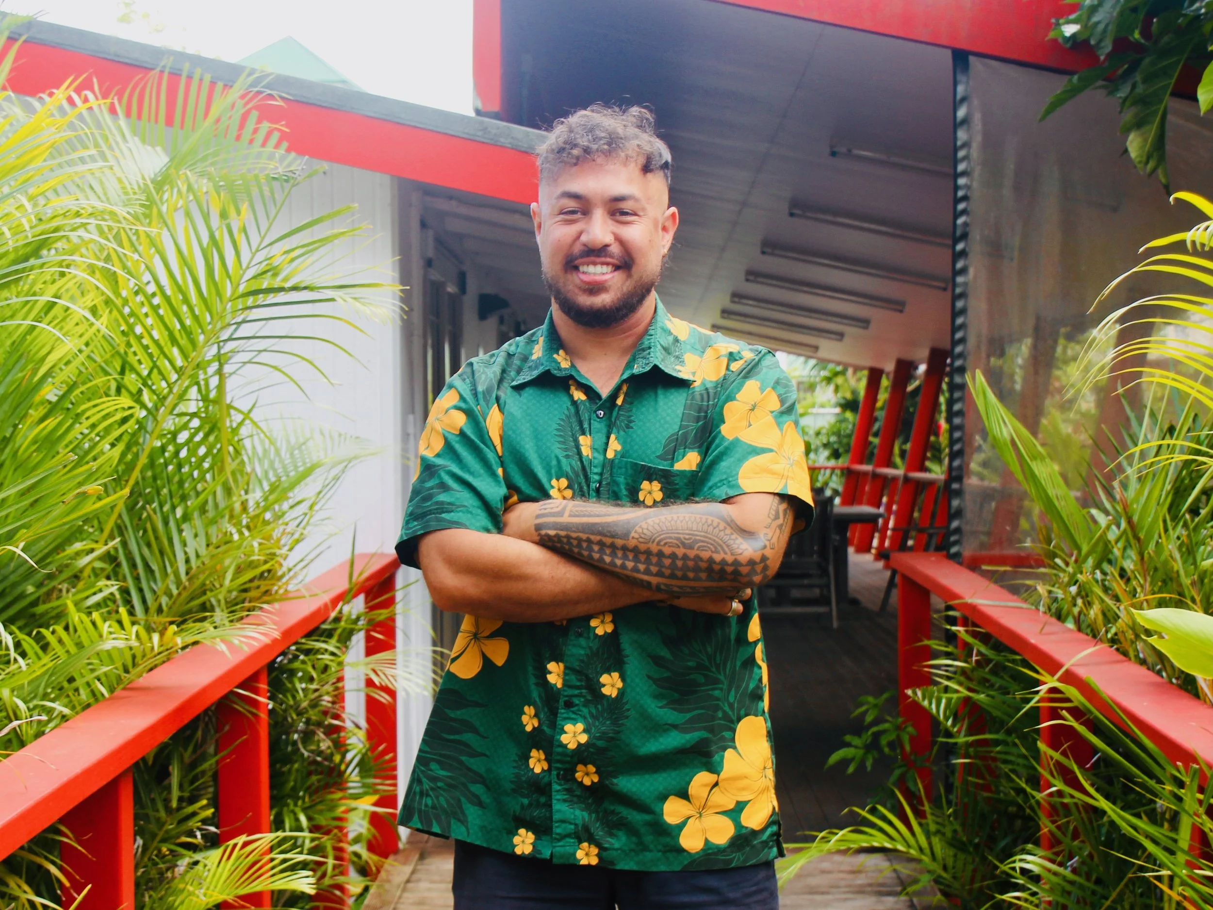 A smiling man with a beard and tattoos on his arm standing on a red bridge surrounded by lush green plants, wearing a green Hawaiian shirt with yellow floral patterns.
