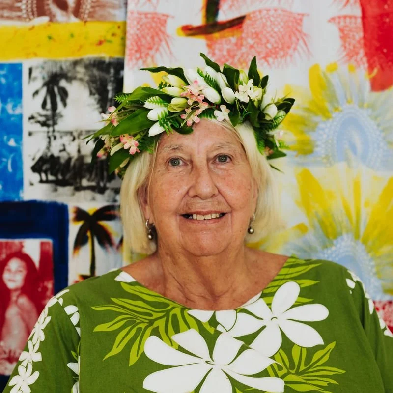 An elderly woman with blonde hair wearing a green dress with white floral pattern and a flower crown with white and pink flowers. Background features colorful, tropical-themed artwork.