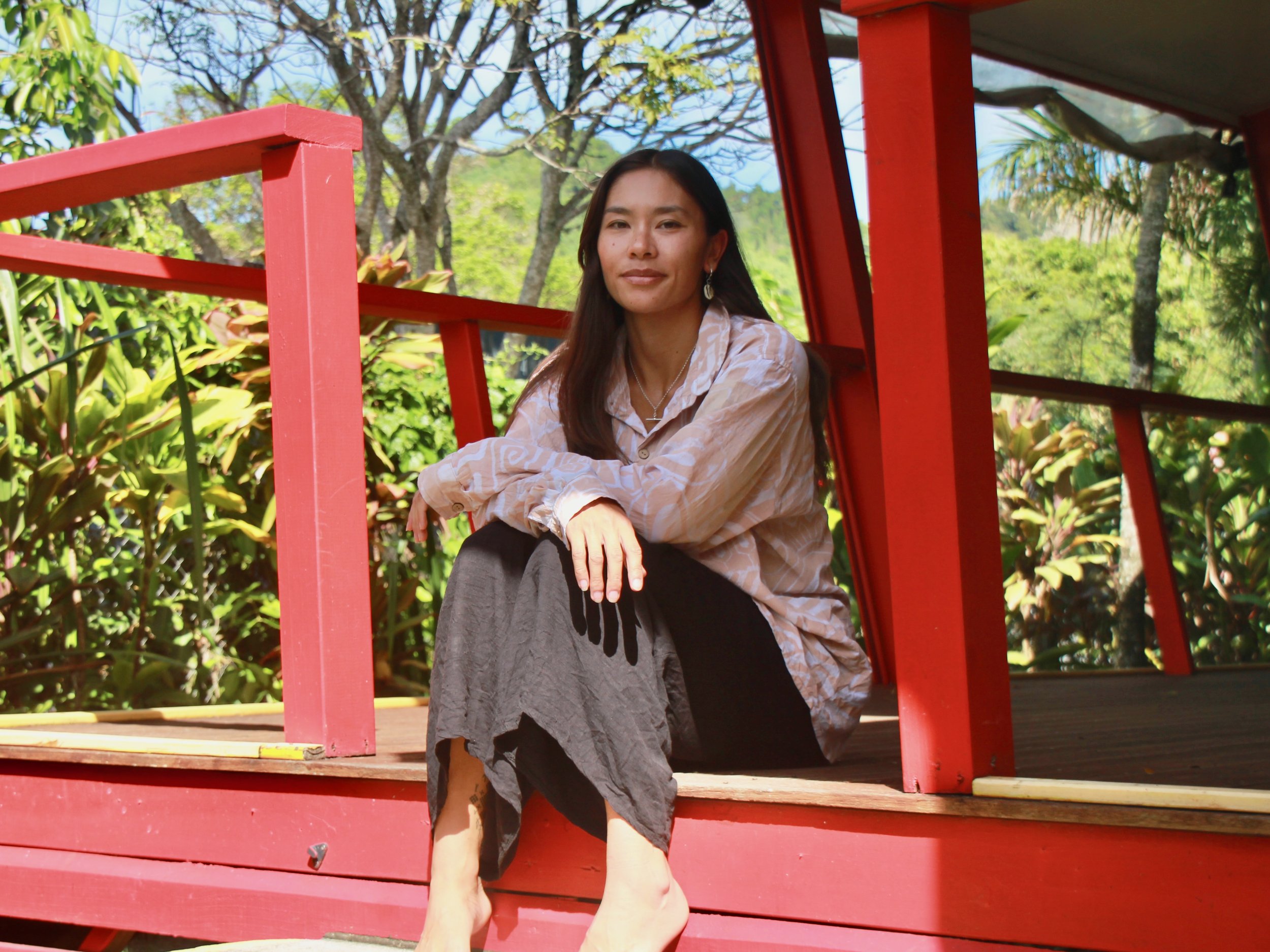 A woman sitting on a wooden porch of a red building surrounded by greenery and trees during the daytime.