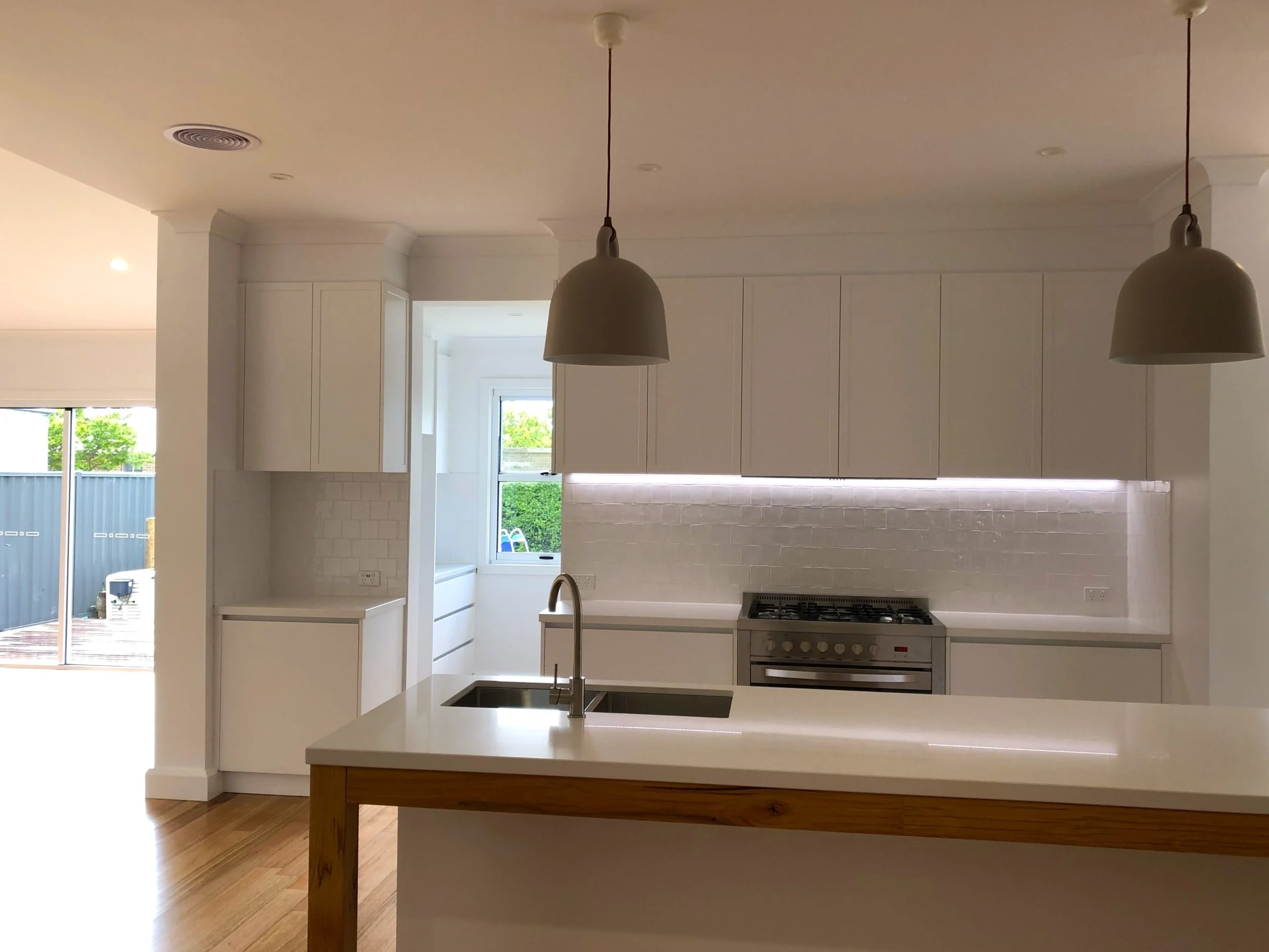 A renovated kitchen with white cabinetry, stone benchtop, timber frame, and two hanging pendant lights over an island with a built-in sink and mixer tap. Tiled Splashback. A sliding glass door leads outside, revealing a backyard with greenery.