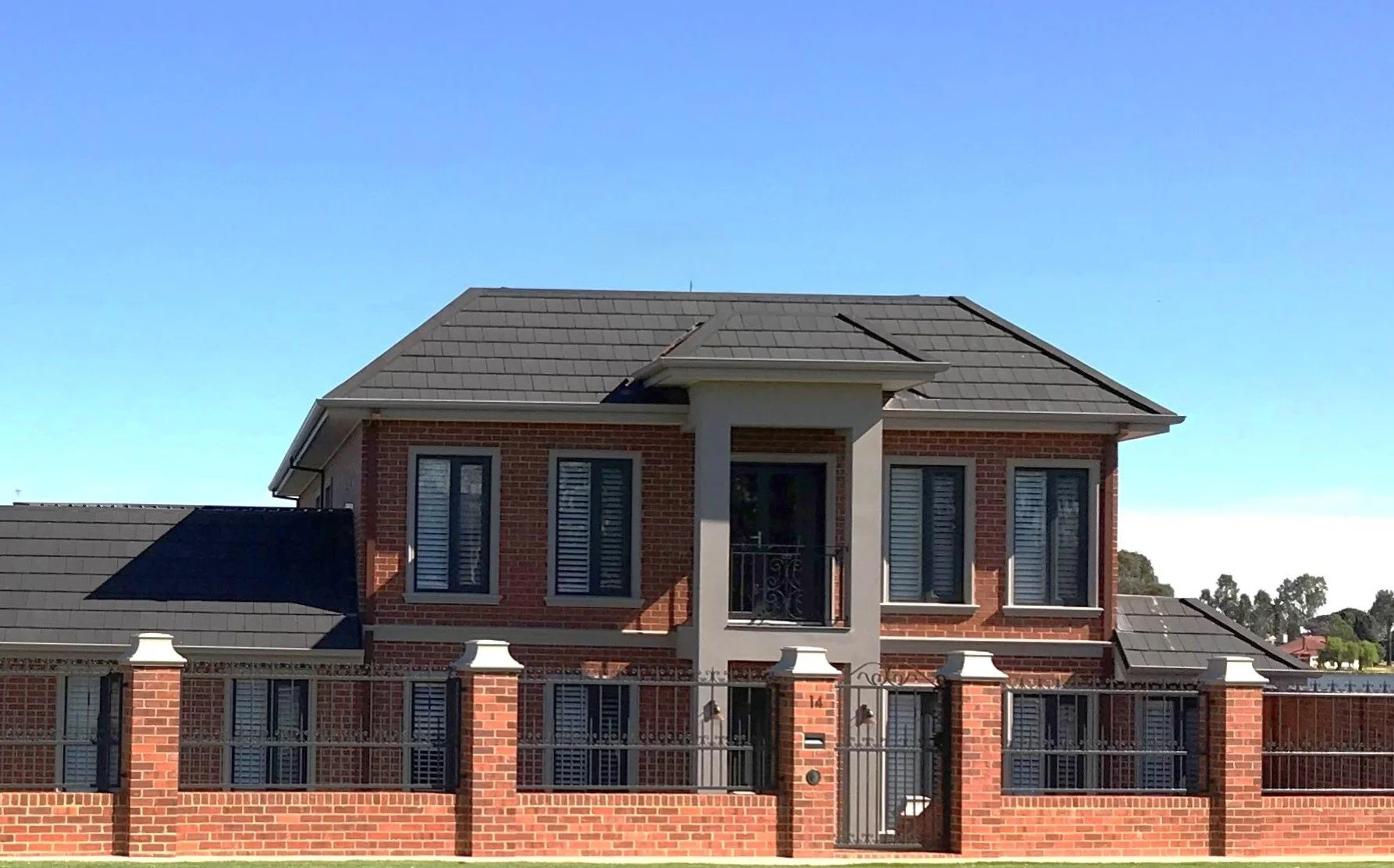 A two-story French Provincial Style brick house with a slate tiled roof, black wrought iron balcony, white shutters, and a brick fence with black wrought iron gate. 