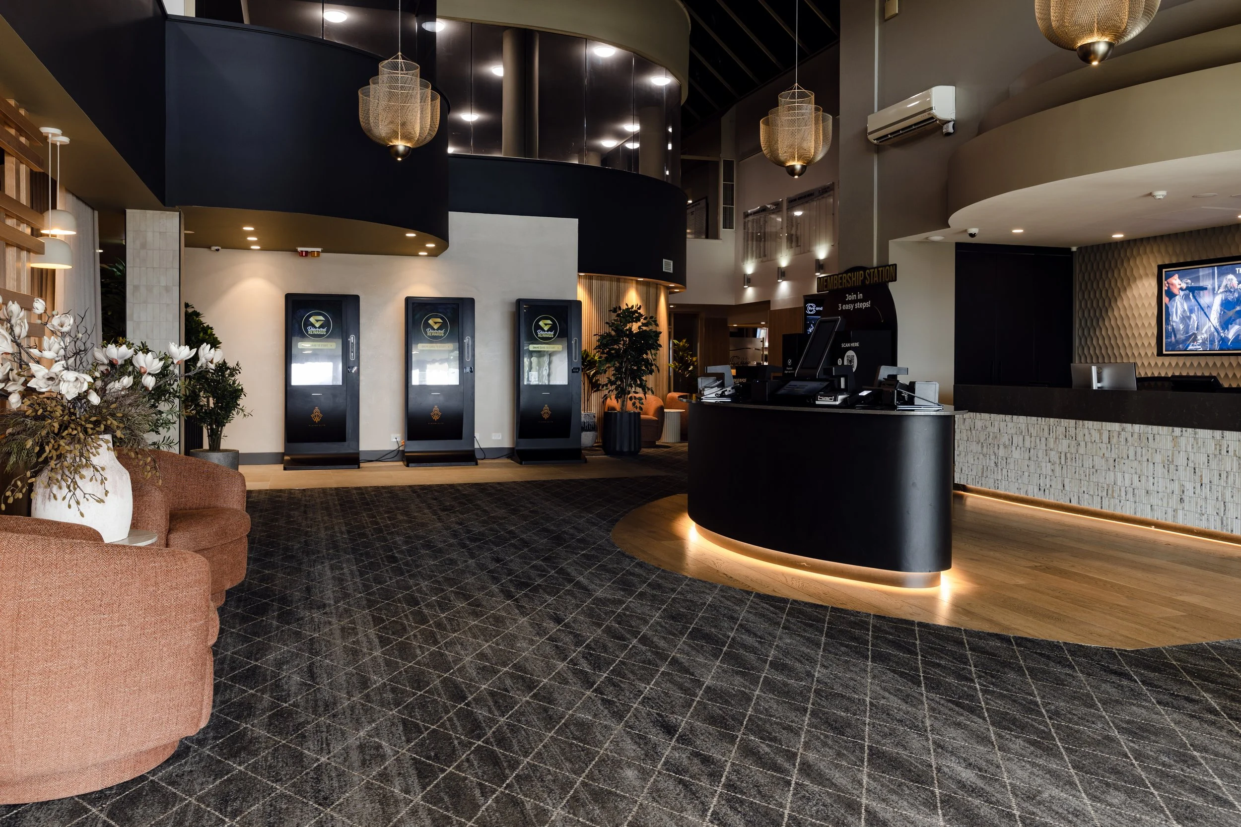 Club Corowa renovation of reception area with black curved reception desk, orange seating, potted plants, a large television screen. Decorative hanging lights and ceiling. Designed by Verity Rhodes Interiors.