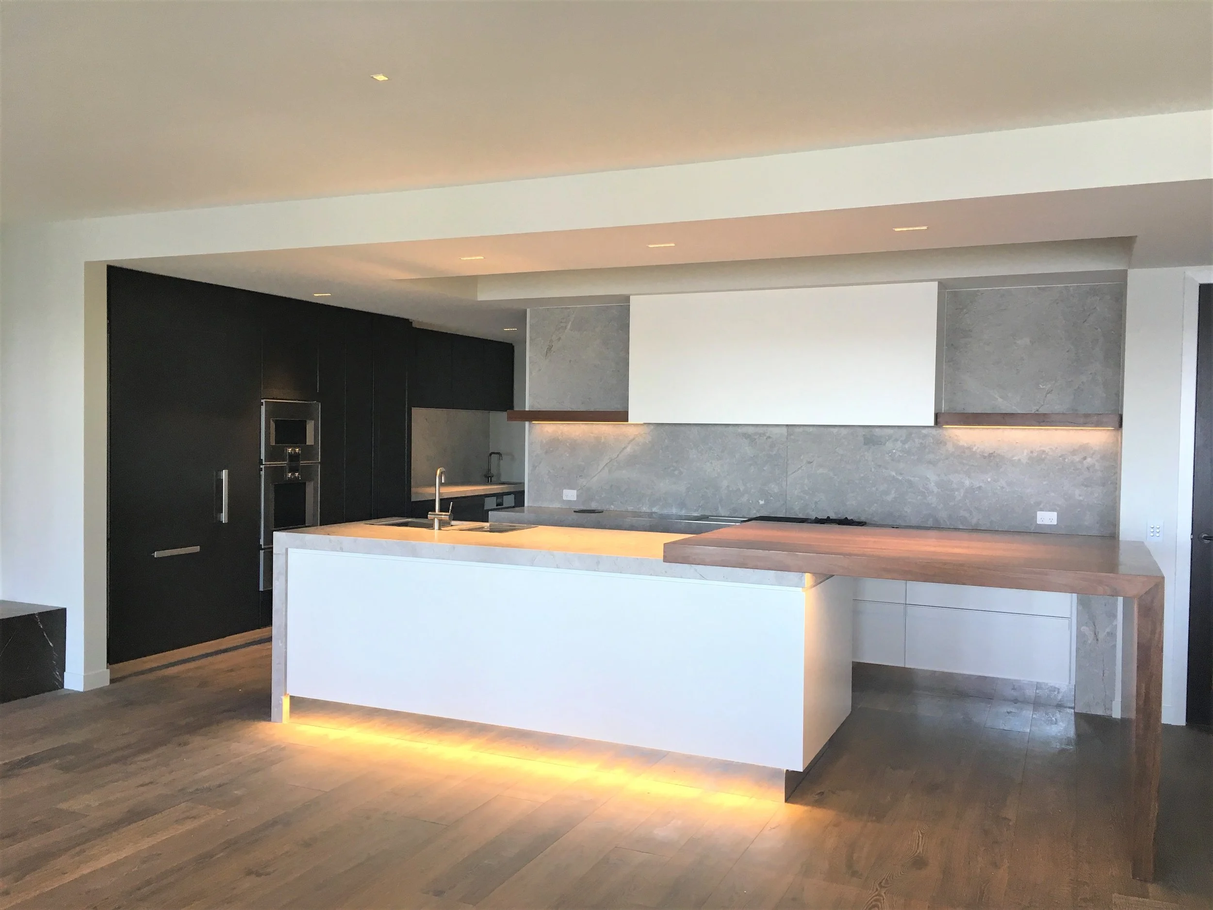 Renovated modern kitchen with black cabinets, white island, wooden flooring, and gray tiled walls with warm ambient lighting.