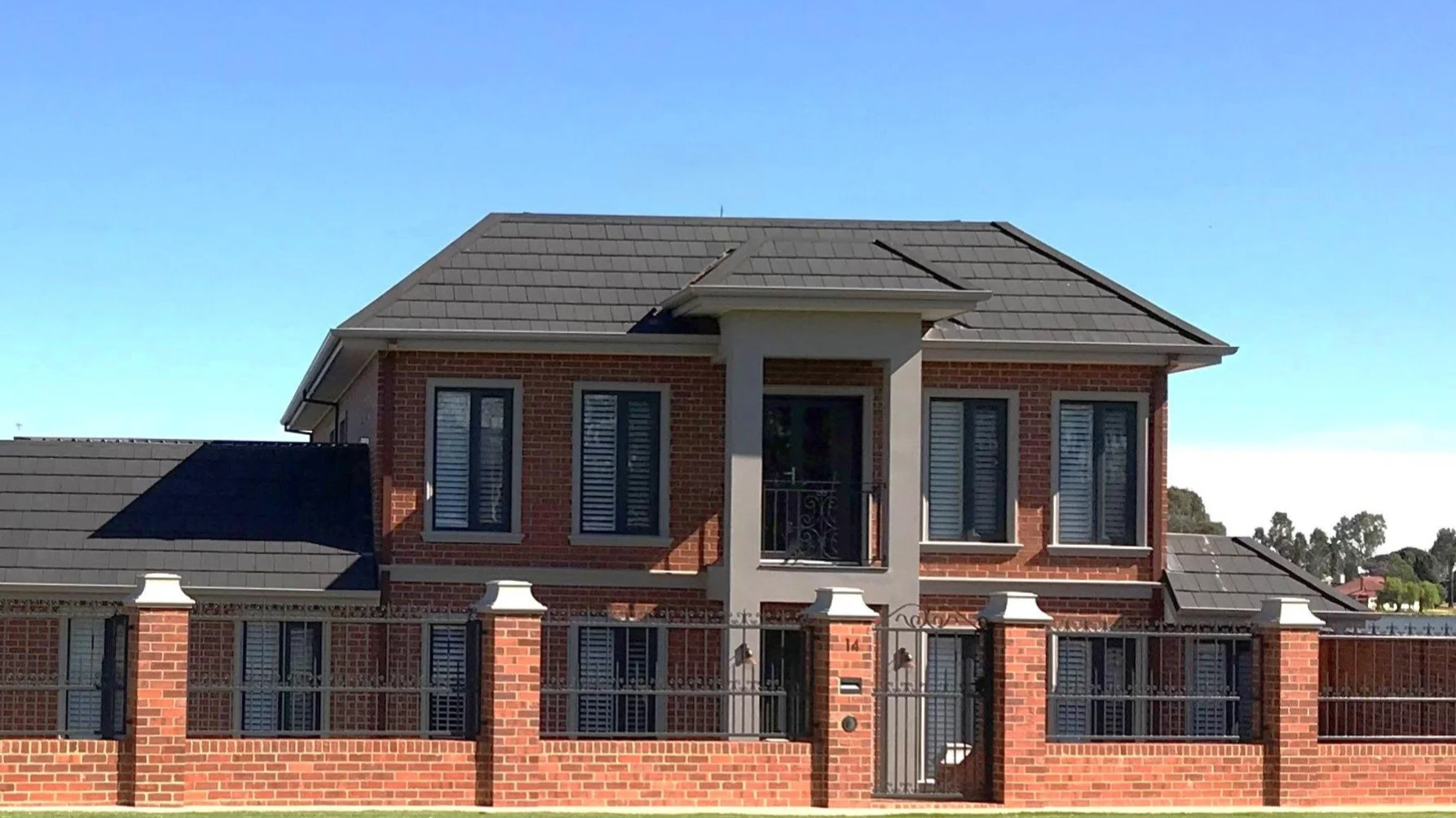 A two-story red brick French Provincial house with black shuttered windows, a gray roof, and a small balcony in front of the middle window on the upper floor. The house is enclosed by a brick and wrought iron fence and gate.