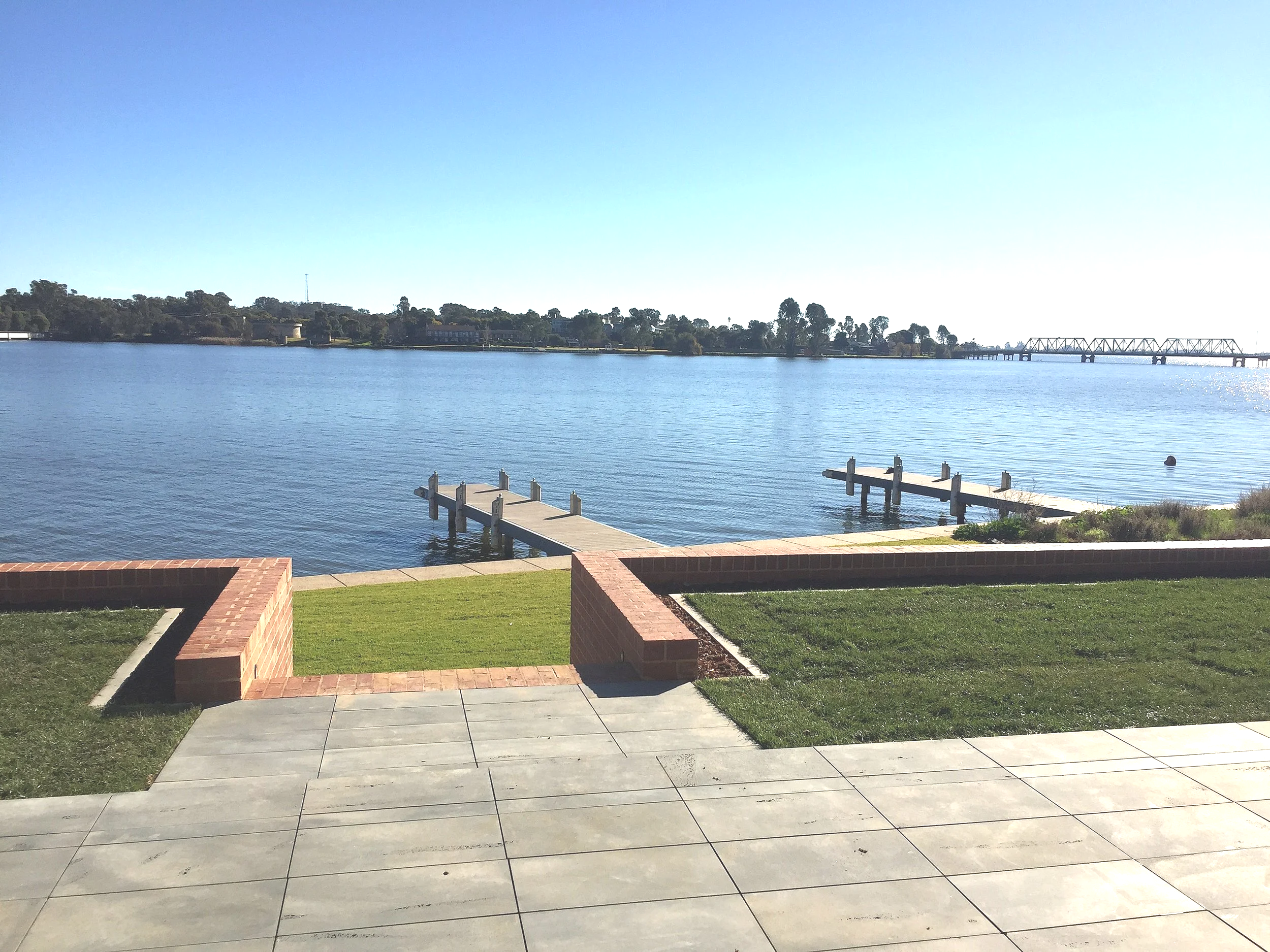 View of Lake Mulwala with a small dock in the foreground, trees along the shore, the Lake Mulwala bridge in the distance, and a clear blue sky.