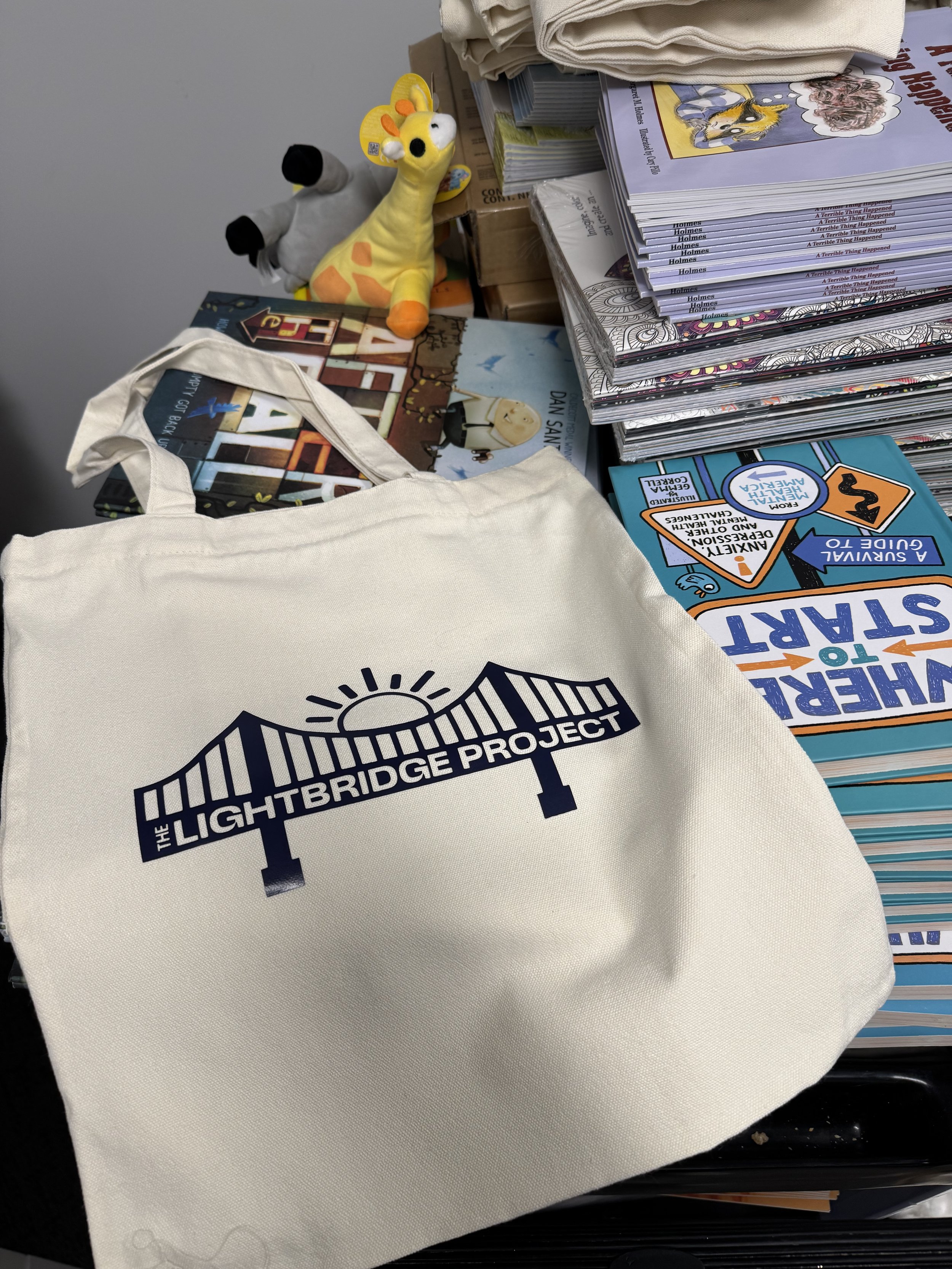 A tote bag with the Lightbridge Project logo on it surrounded by stacks of books, a plush giraffe toy, a plush dog toy, and some children's booklets on a table.