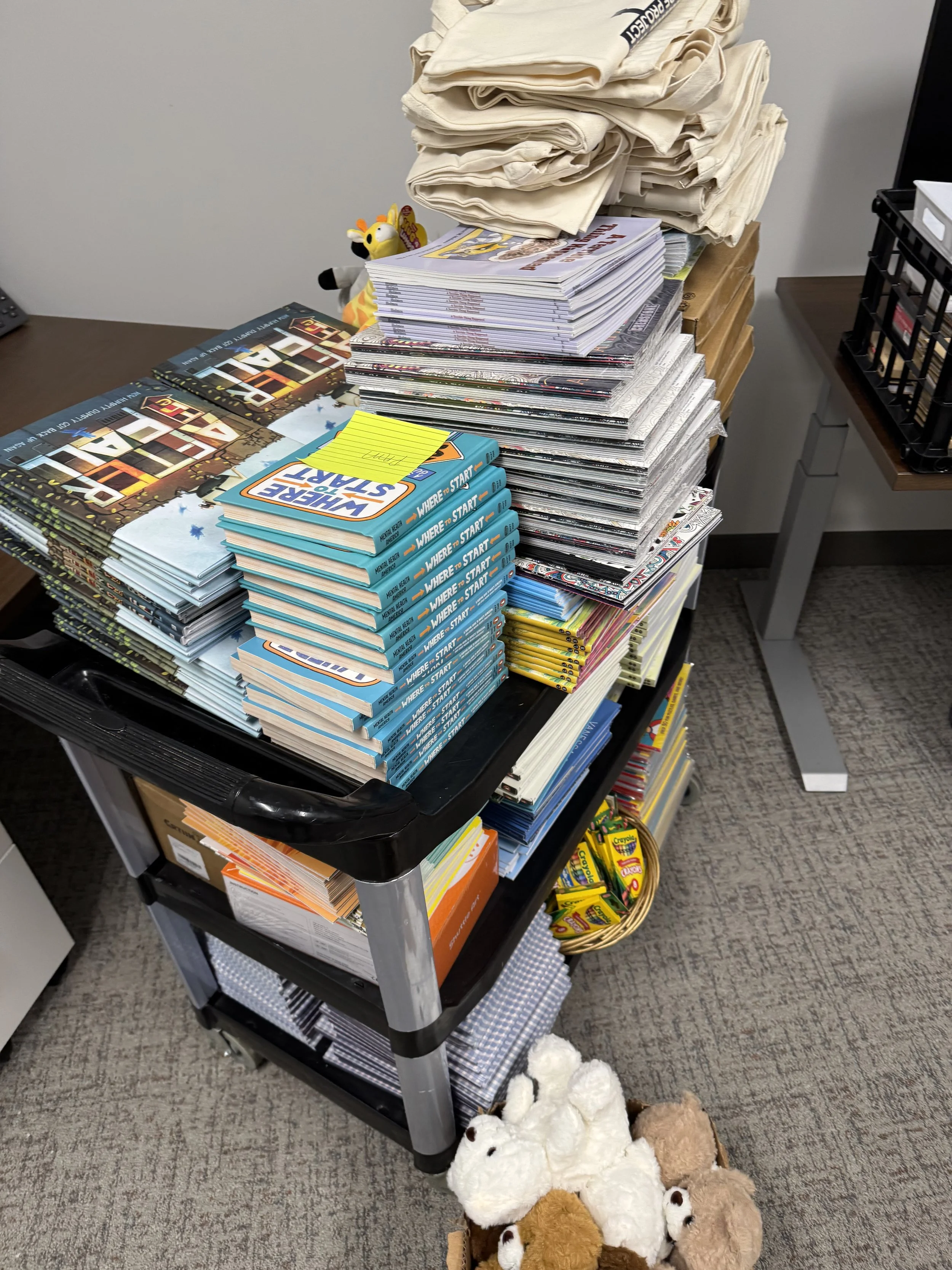 A black rolling cart filled with stacks of textbooks, pamphlets, and paper organizations, with stuffed animals on the floor in front and a desk on the side.