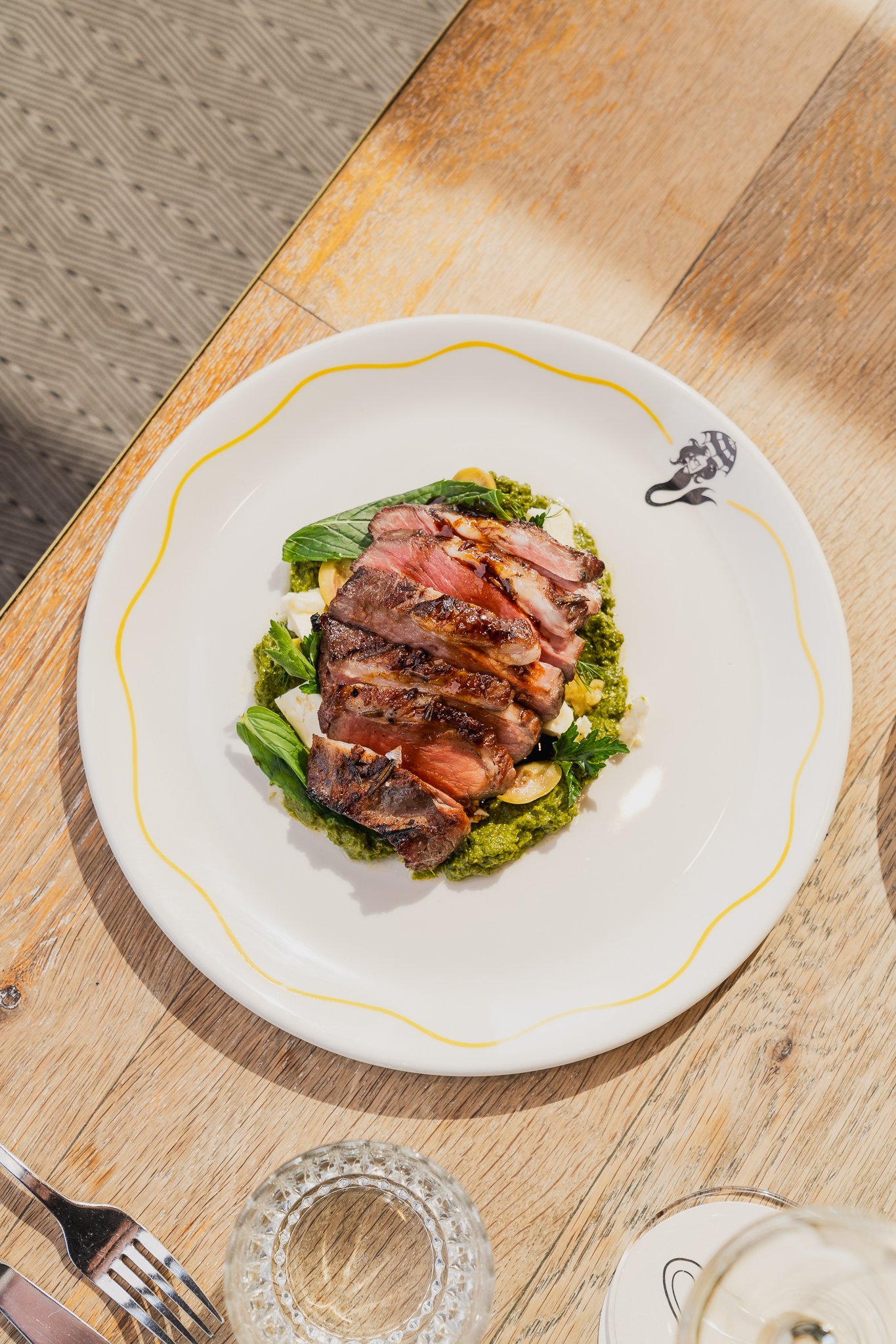 A white plate with a small border featuring a black cartoon character holding an umbrella, round salad greens, slices of steak, and a green pesto sauce on a wooden table. Glasses and utensils are also on the table.