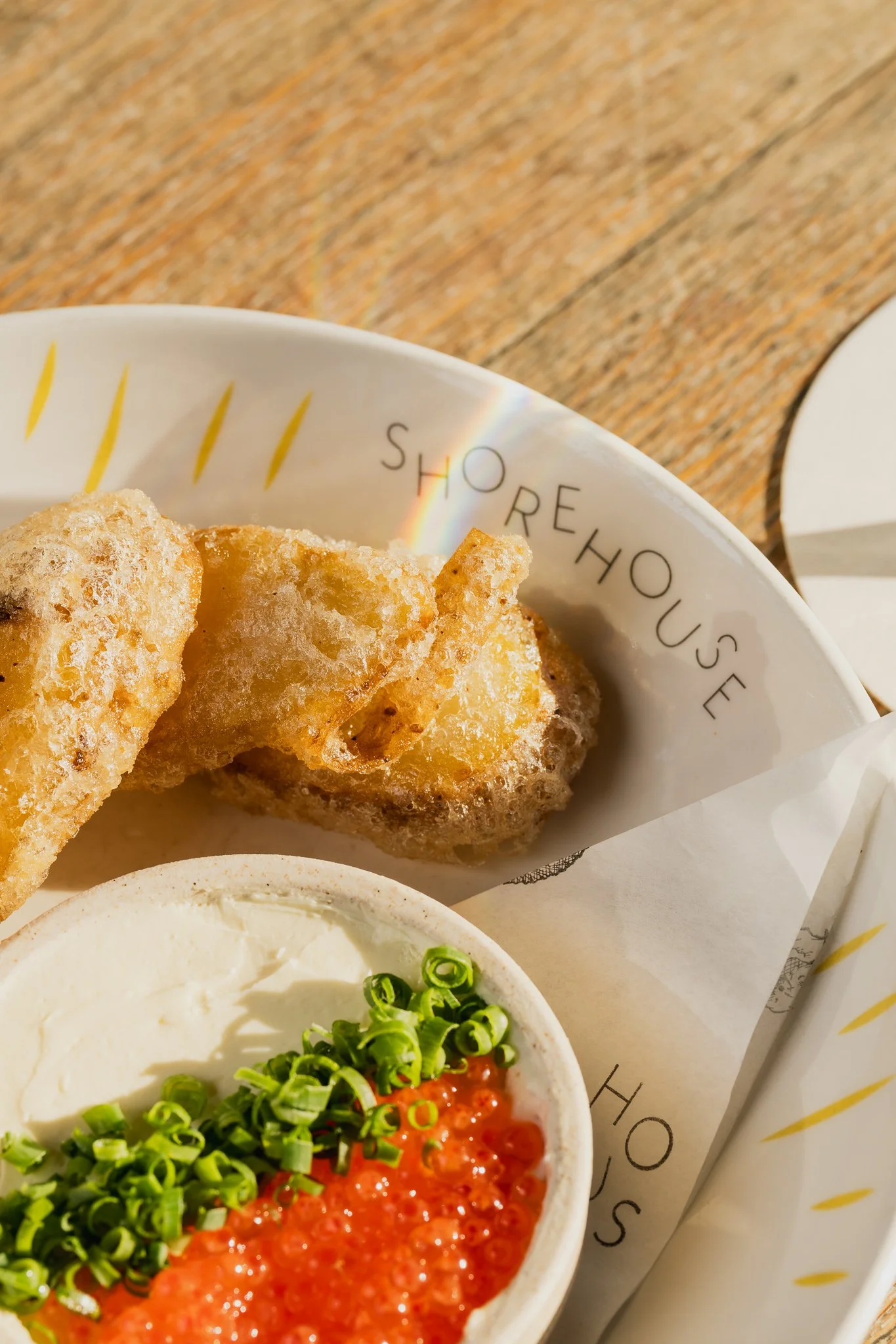 Close-up of fried food served in a paper bowl with yellow and black text, and a bowl of dip with green onions garnish.