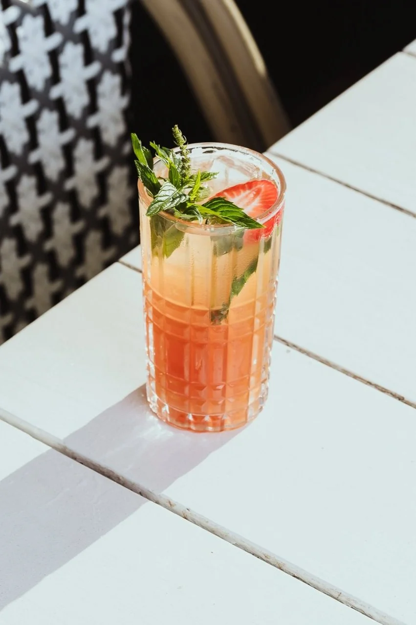 A pink-colored cocktail in a textured glass, garnished with fresh strawberries, mint leaves, and a sprig of herbs, placed on a white outdoor table.