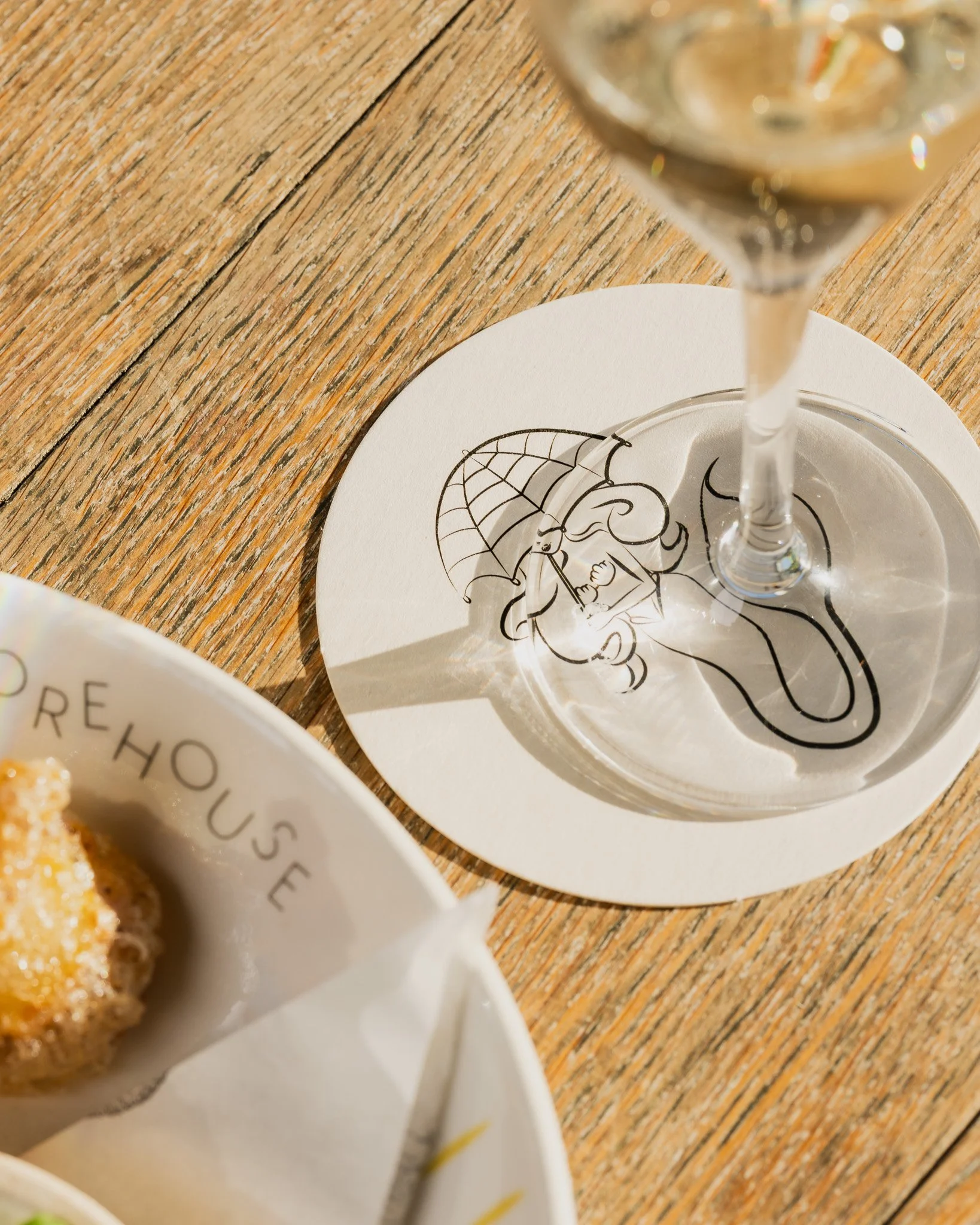 A table with a glass of white wine, a decorative paper coaster featuring an illustration of a girl with an umbrella, and a plate of cookies on a wooden surface.