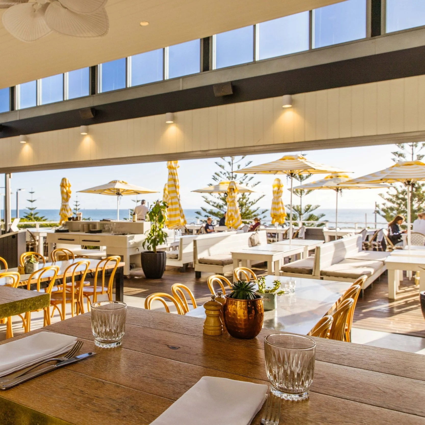 Indoor restaurant with wooden dining tables and chairs, overlooking an outdoor patio with yellow striped umbrellas, trees, and the ocean in the background.