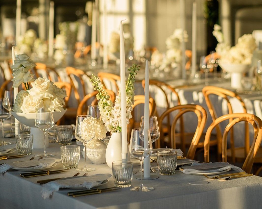 Elegant banquet table decorated with white floral arrangements and tall white candles, set for a formal event with glassware, plates, and gold utensils in a sunlit room.