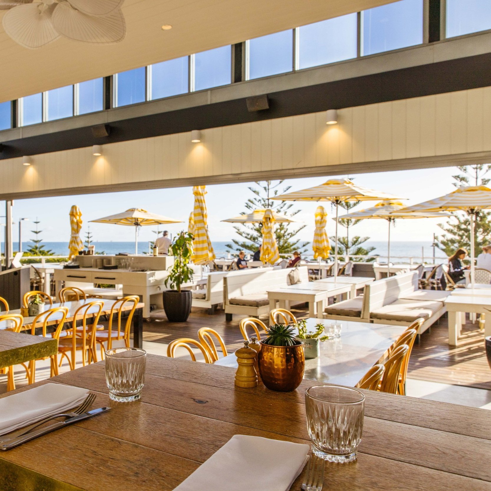 Indoor restaurant with a view of outdoor patio, yellow chairs, white tables, tropical plants, and umbrellas near the beach.