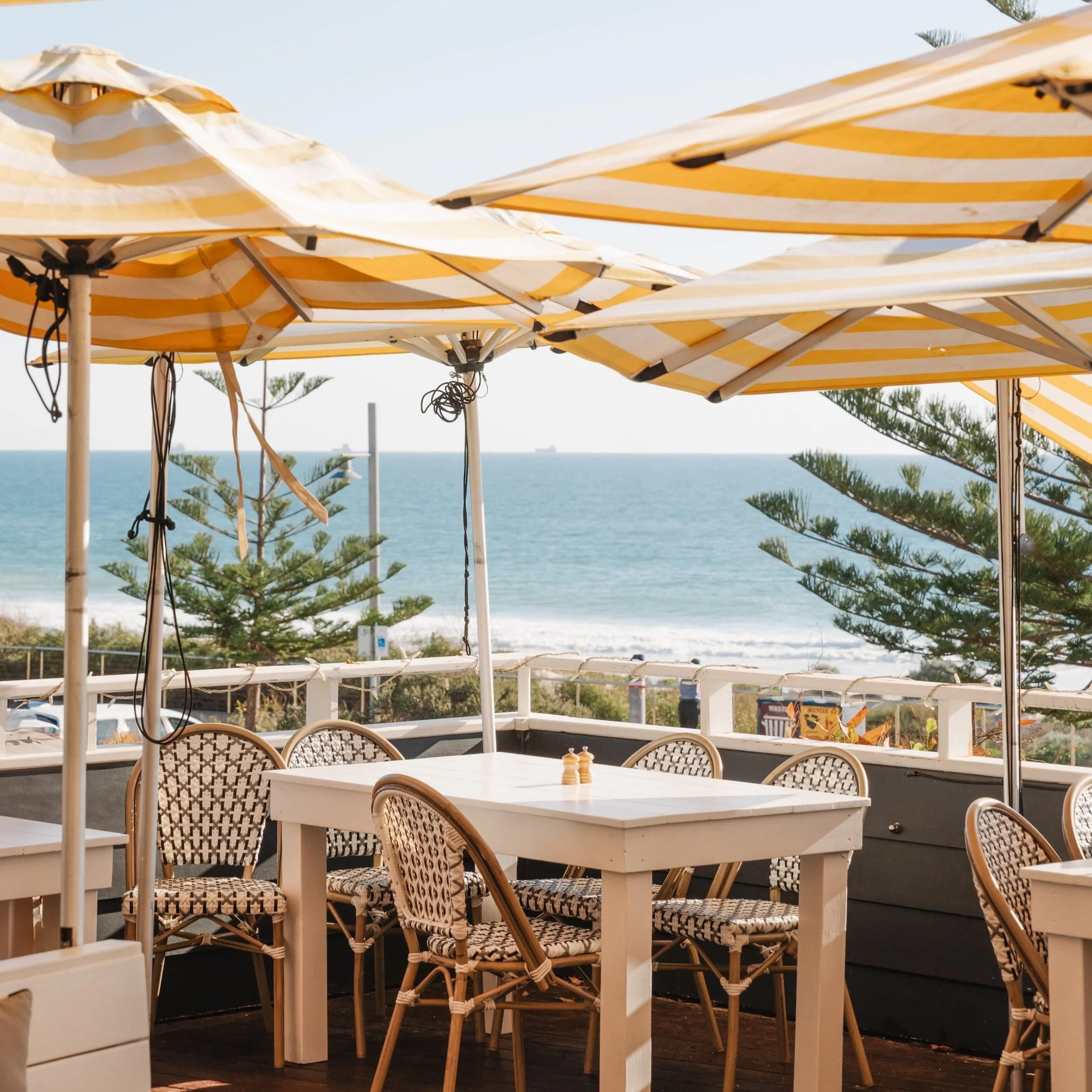 Beachside restaurant with white tables and woven chairs under large yellow and white striped umbrellas, overlooking the ocean with ships in the distance.