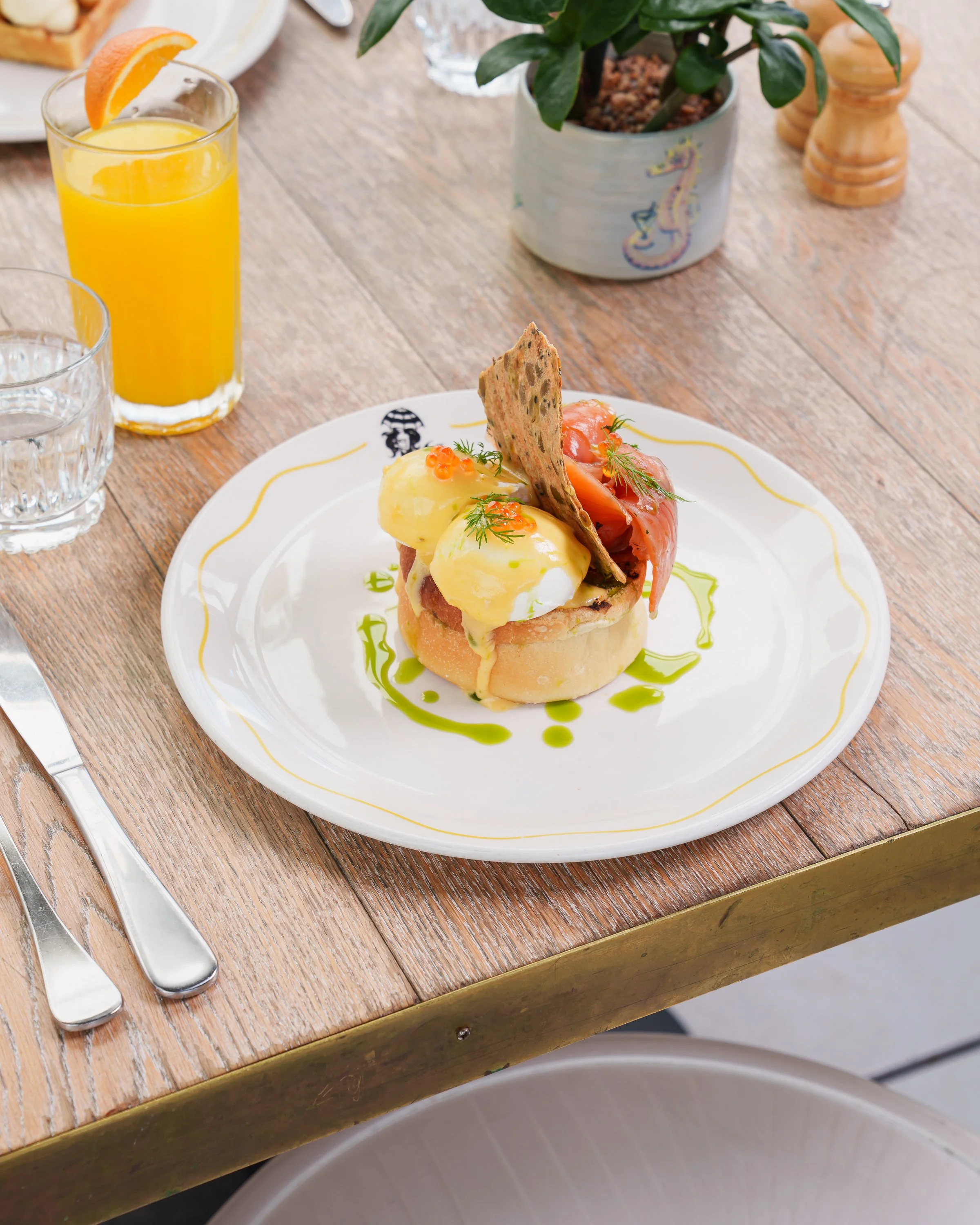 A plate of eggs Benedict with smoked salmon, garnished with dill and small orange fish roe, served with a side of smoked salmon and a crispy cracker, on a white plate with a yellow and green design. There is a glass of orange juice with a slice of orange, a glass of water, a plant in a decorative pot, and cutlery on a wooden table.