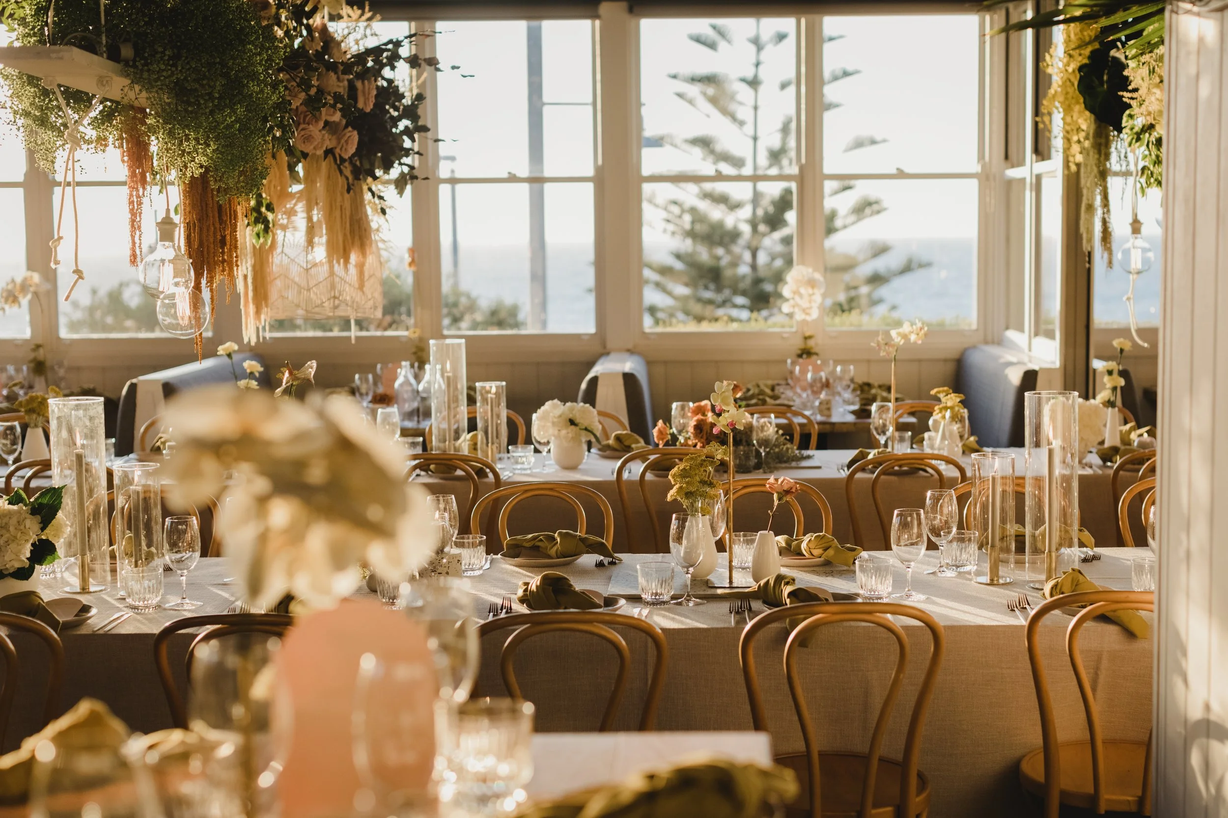 Elegant indoor event space decorated with floral arrangements, glass vases, and table settings, overlooking a scenic seaside view through large windows.
