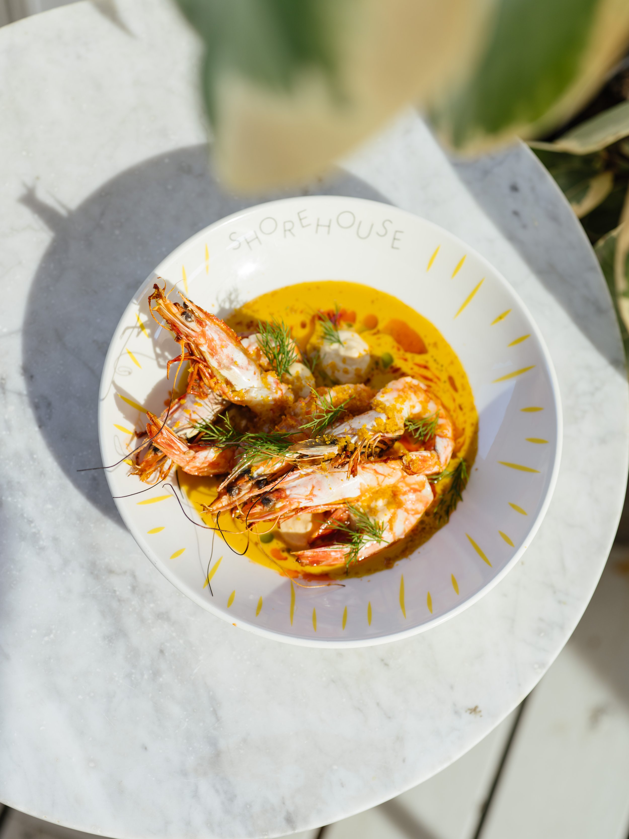 A bowl of shrimp and seafood stew garnished with herbs, served on a white marble table outdoors.