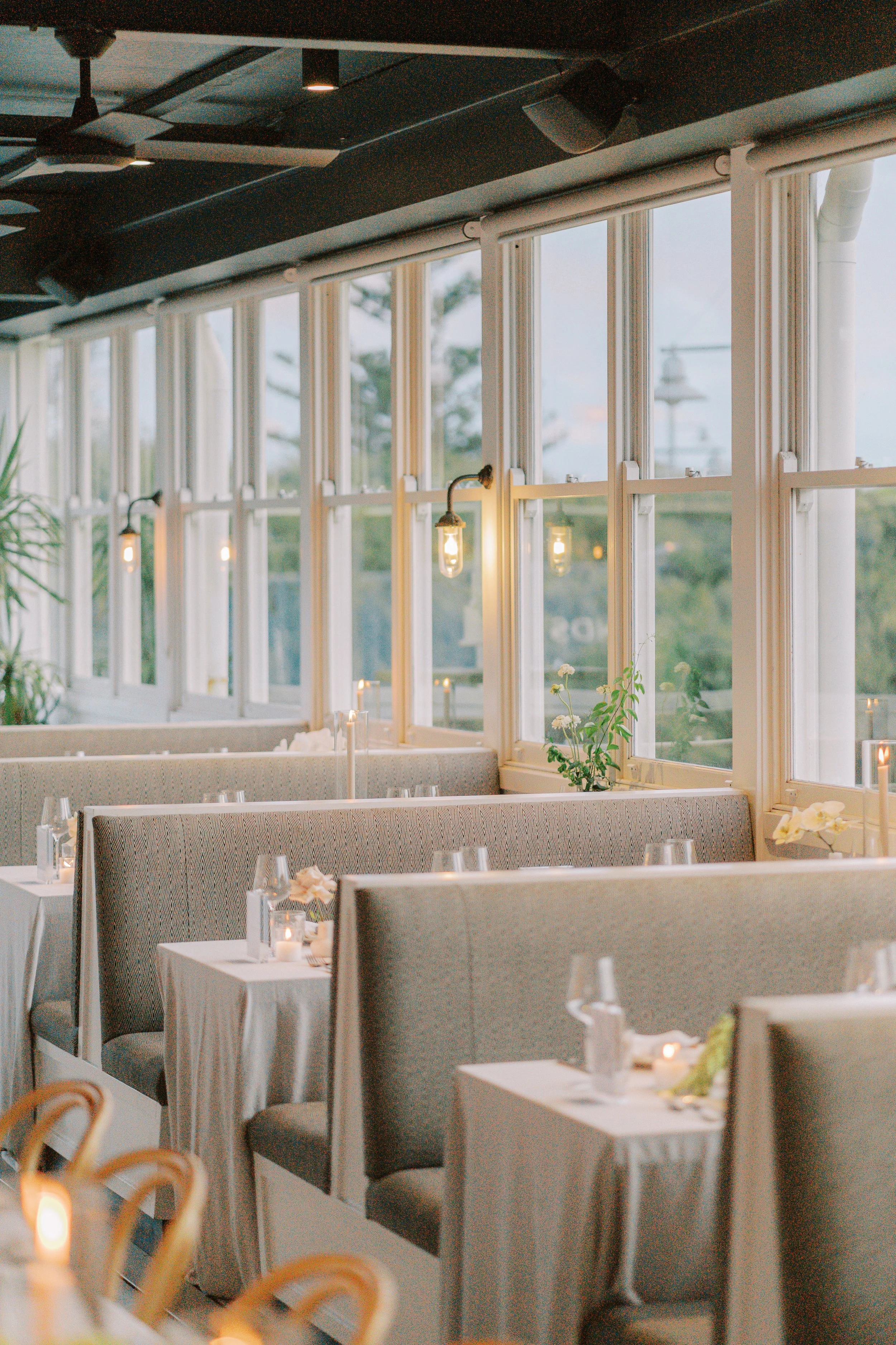 Interior of a restaurant with large windows, decorated tables with white tablecloths, small candles, flowers, and place settings, and wall-mounted light fixtures.