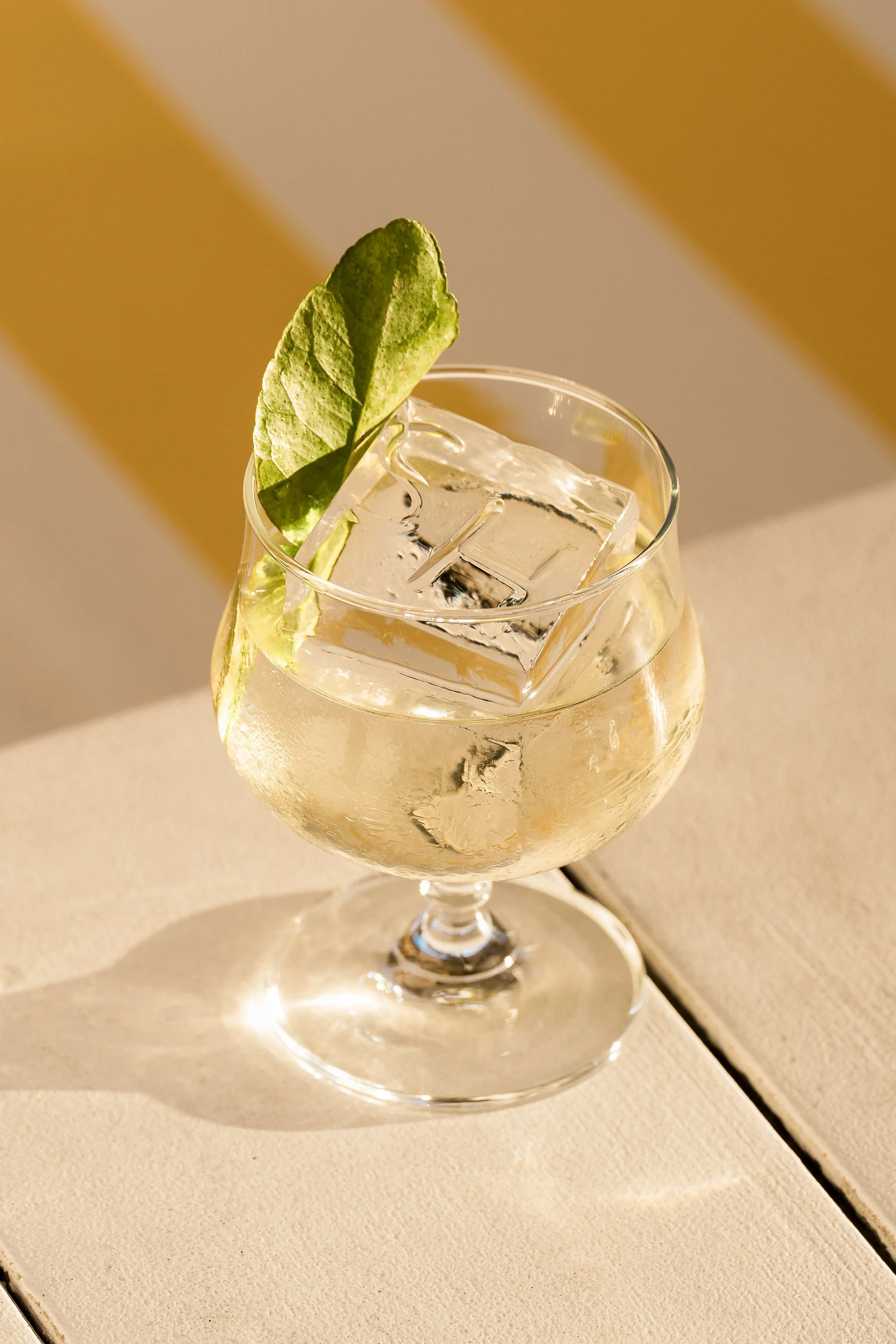 A glass of clear beverage with ice cubes, garnished with a green mint leaf, on a white surface.
