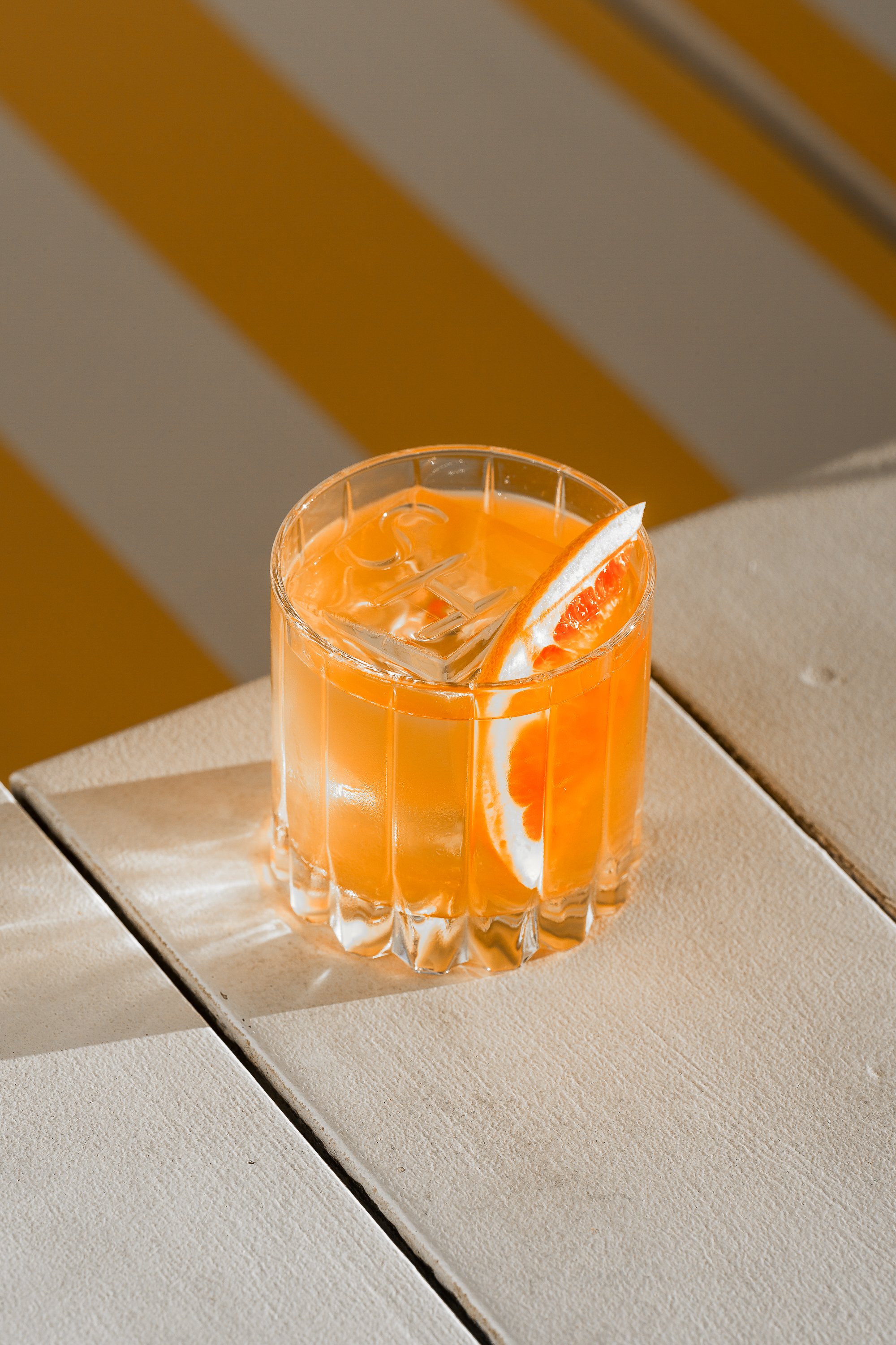 A glass of orange juice with ice and a slice of orange on the rim, placed on a beige surface with a background of yellow and gray diagonal stripes.