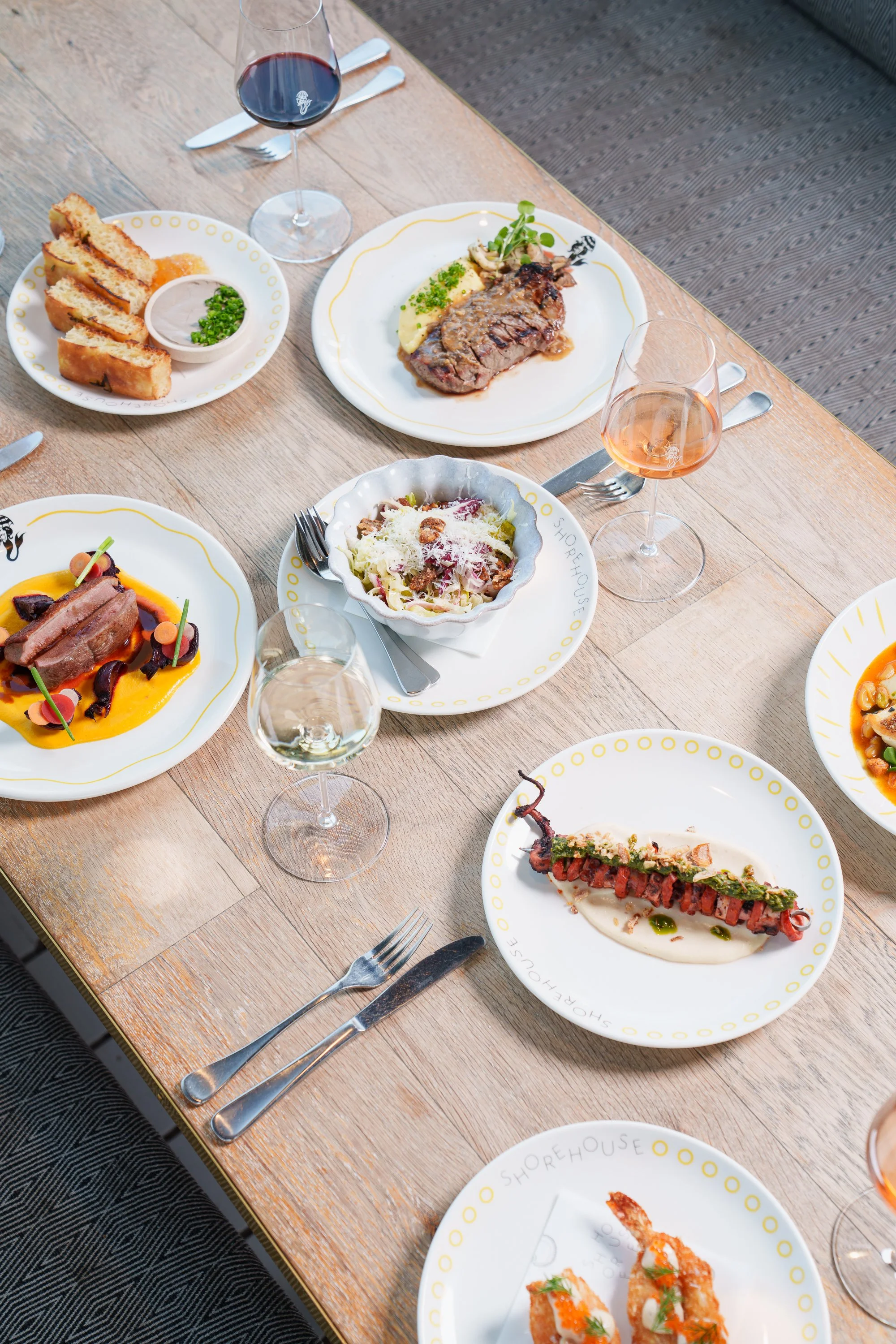 A dining table with various plated dishes and glasses of wine, set with cutlery in a restaurant or cafe.