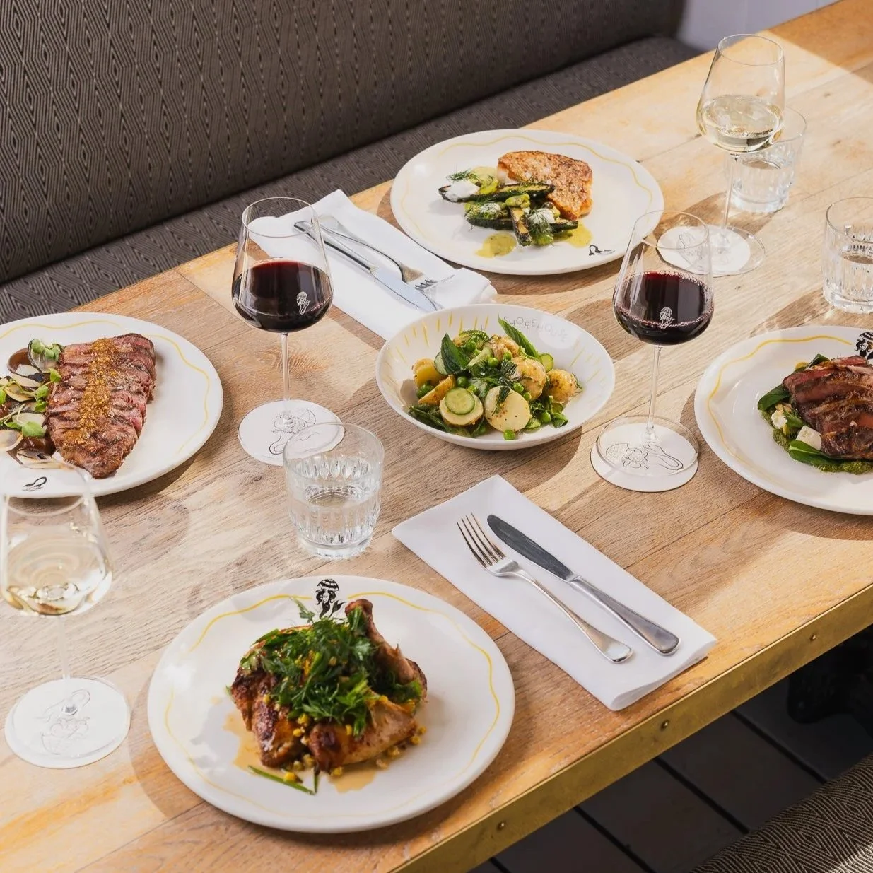 A wooden dining table set with plates of cooked meats, salads, wine glasses filled with red wine, and water glasses, with silverware and napkins.