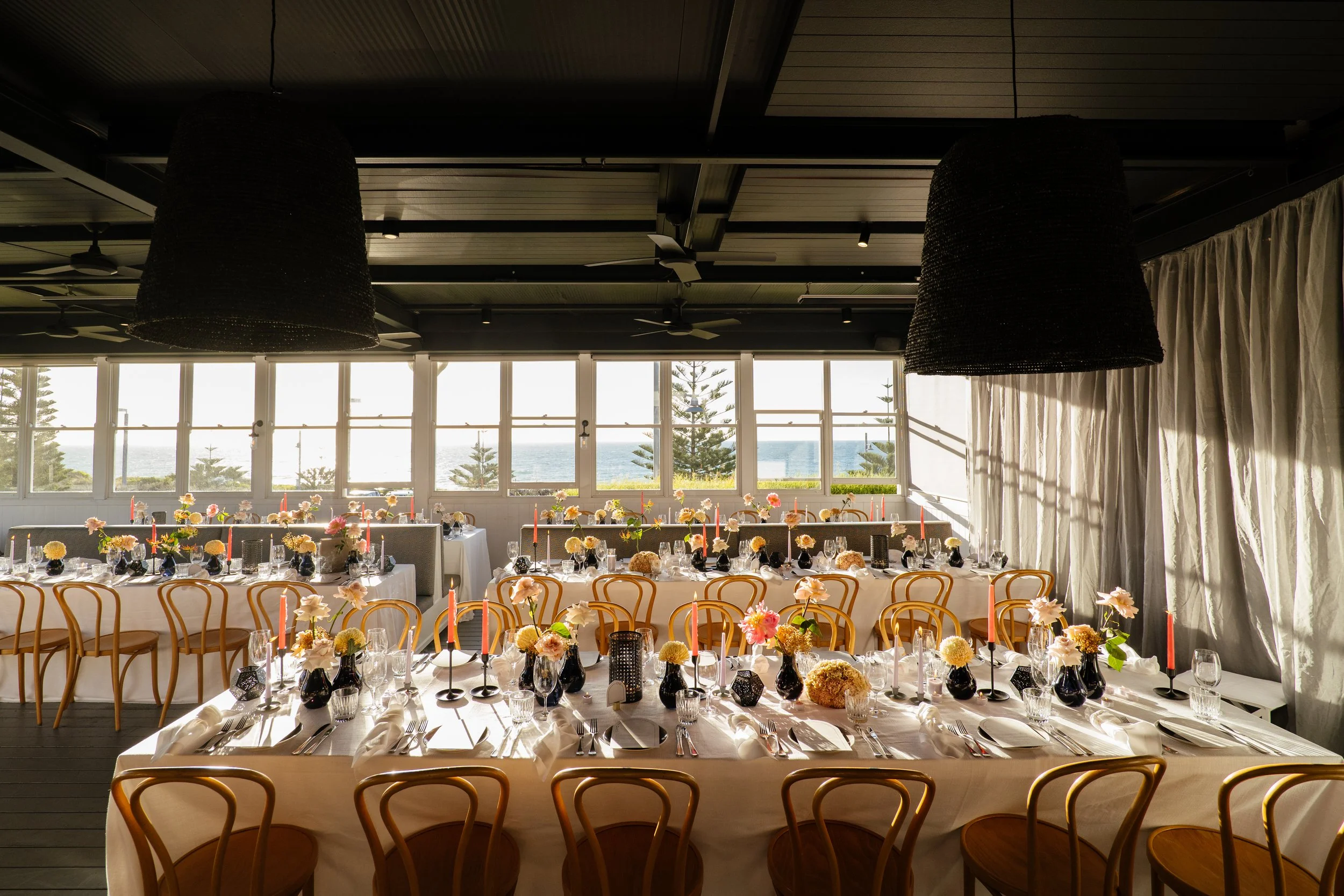 Elegant indoor event space with long tables set for a meal, decorated with flowers and candles, large windows revealing a view of trees and the ocean, dark ceiling with large pendant lights, and white curtains.