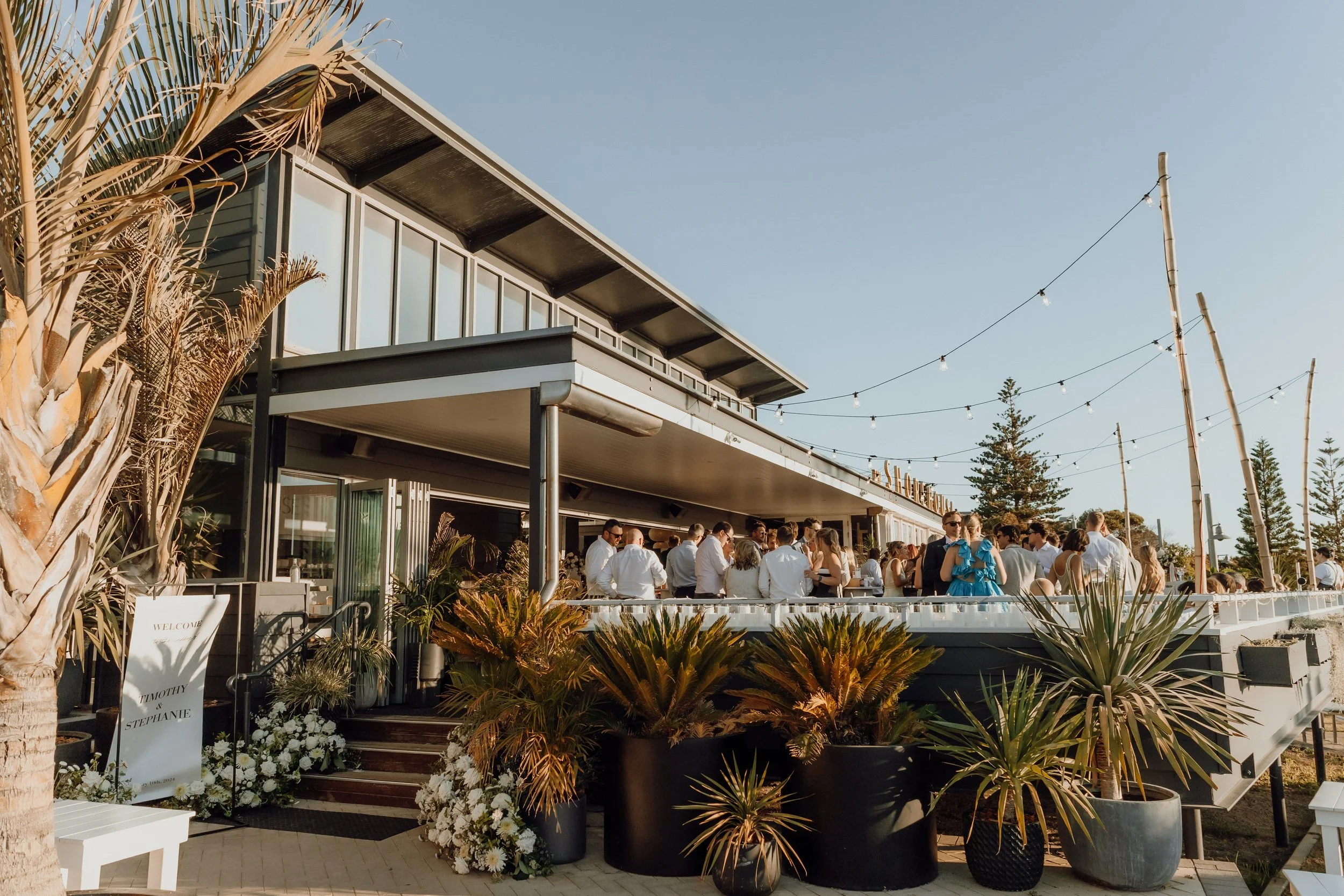 People gathered at an outdoor event on a patio of a modern building with glass windows and string lights, surrounded by tropical plants and trees during daylight.