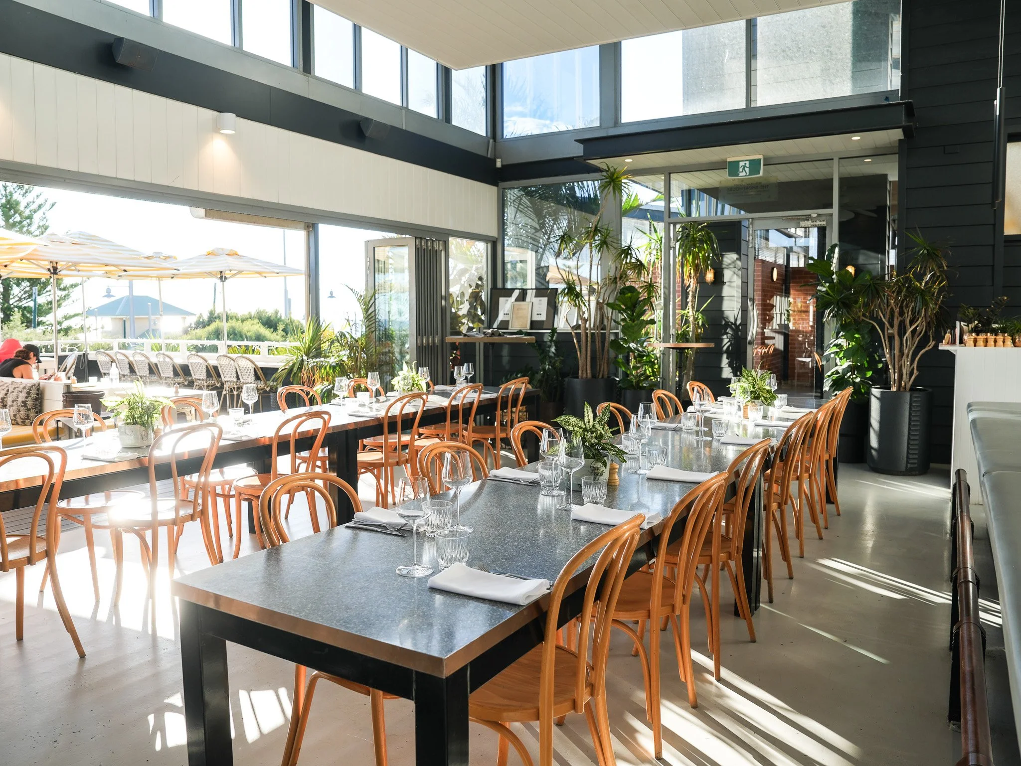 Interior of a modern restaurant with wooden chairs, tables set with glasses and napkins, large floor-to-ceiling windows, and greenery visible outside.