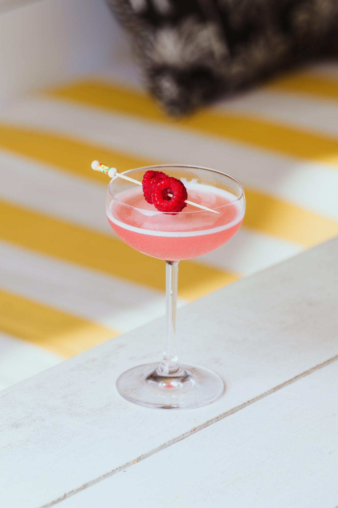 A pink cocktail in a glass with a raspberry garnish on top, placed on a white surface with a yellow and white striped background.