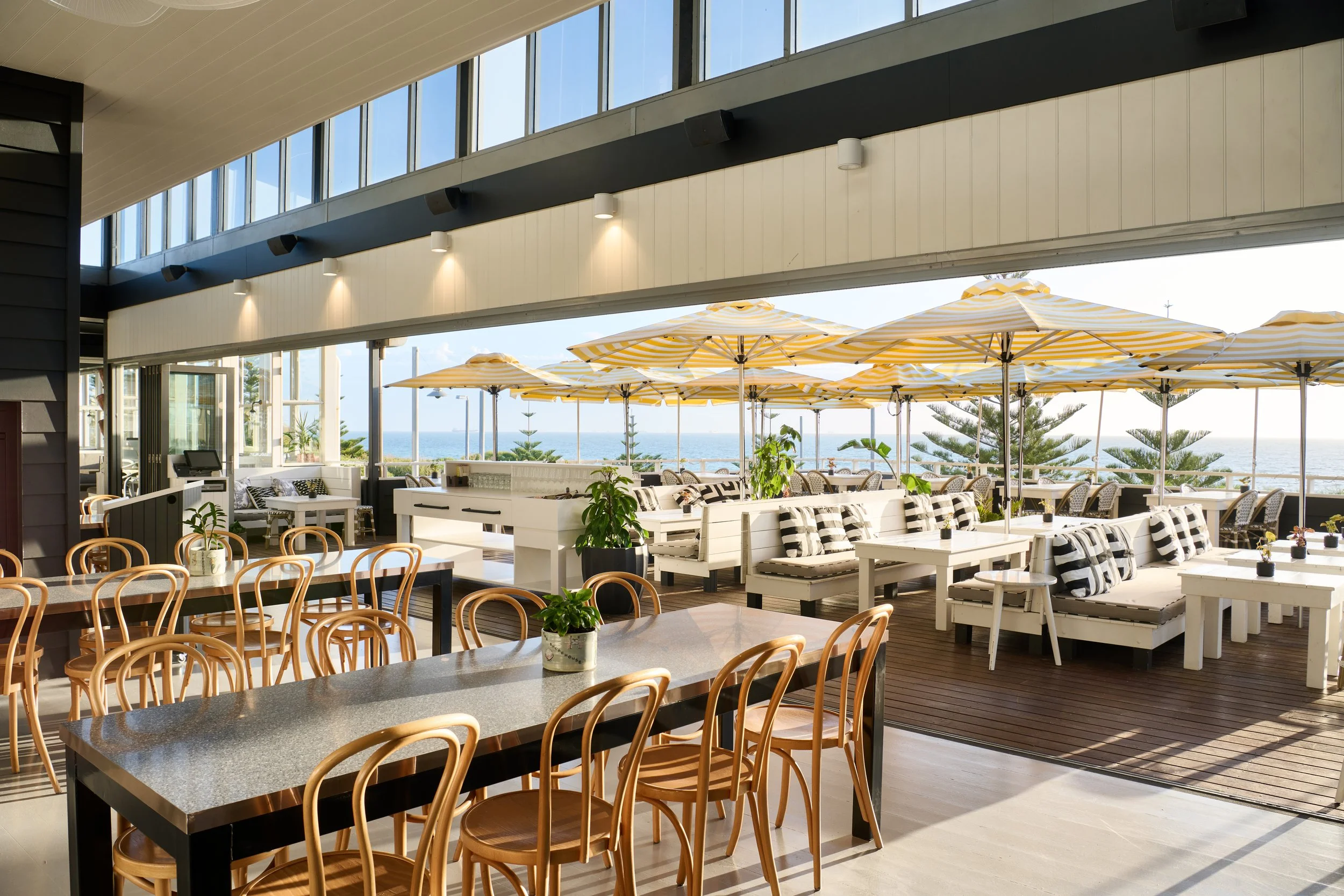 Indoor seating area of a restaurant or cafe overlooking the ocean, with large windows, yellow and white striped umbrellas outside, and a mix of wooden and white upholstered furniture.
