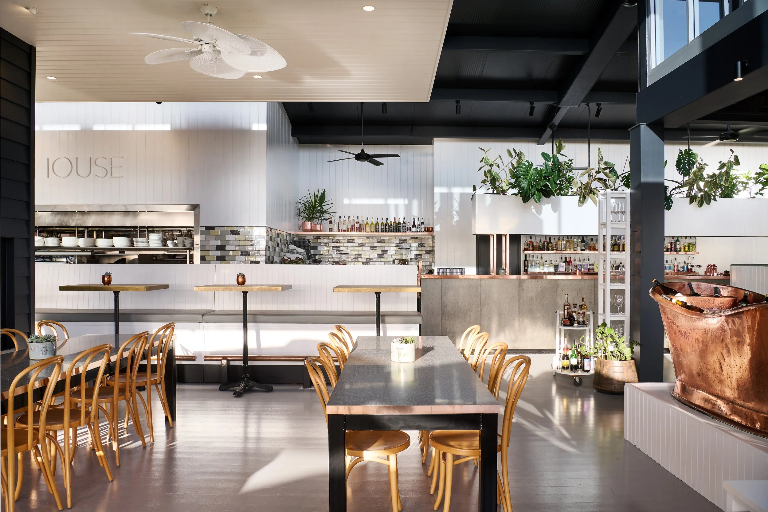 Interior of a modern restaurant with wooden chairs, black tabletops, and green plants. The ceiling has white and black sections with ceiling fans and lighting. Bar area with bottles and plants is visible in the background.