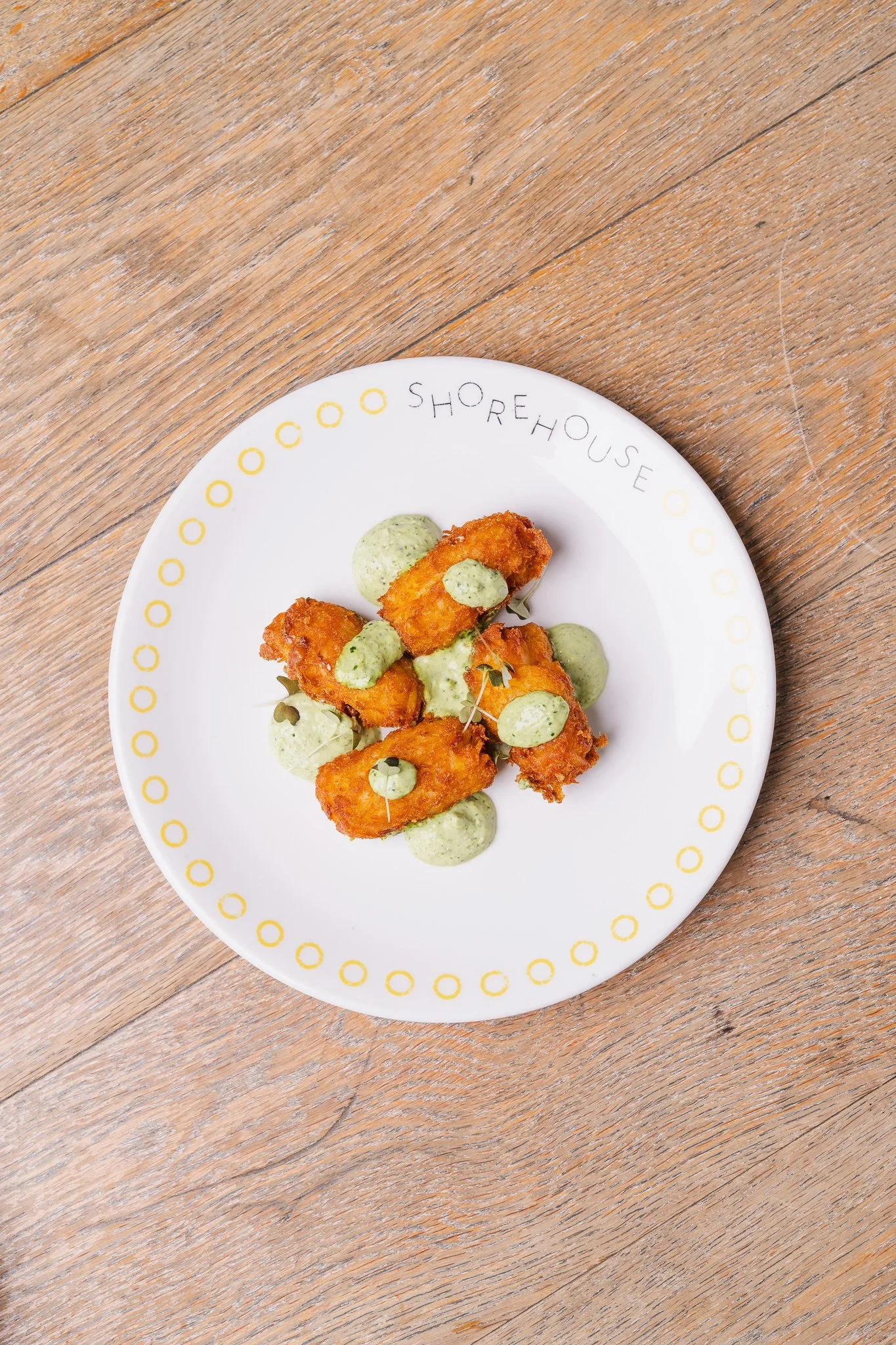A white plate with yellow circle border logo from Shorehouse, containing fried fish pieces topped with green herb sauce, on a wooden table.