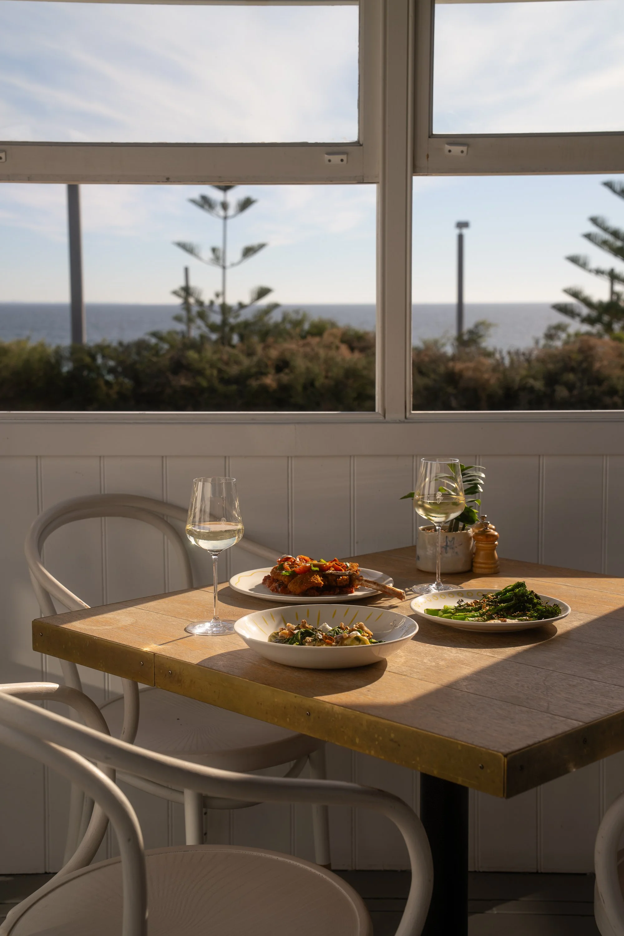 A wooden dining table set with various plates of food, wine glasses, and a small potted plant, with ocean view through open window in the background.