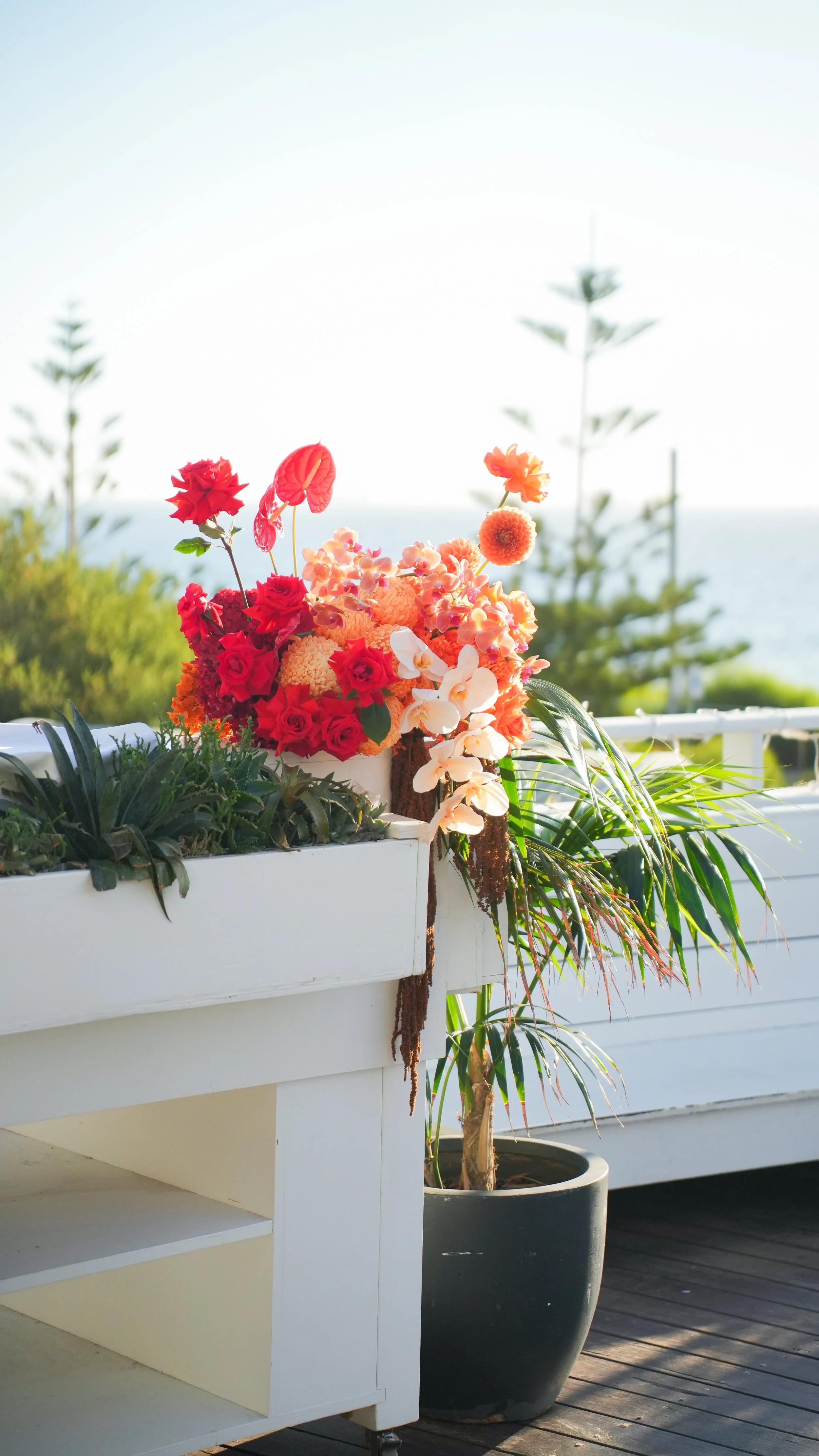 A white outdoor planter filled with various colorful flowers, including red, pink, peach, and white blooms, on a wooden deck with a large black pot beside it, against a backdrop of green trees, a white railing, and a body of water under a clear sky.