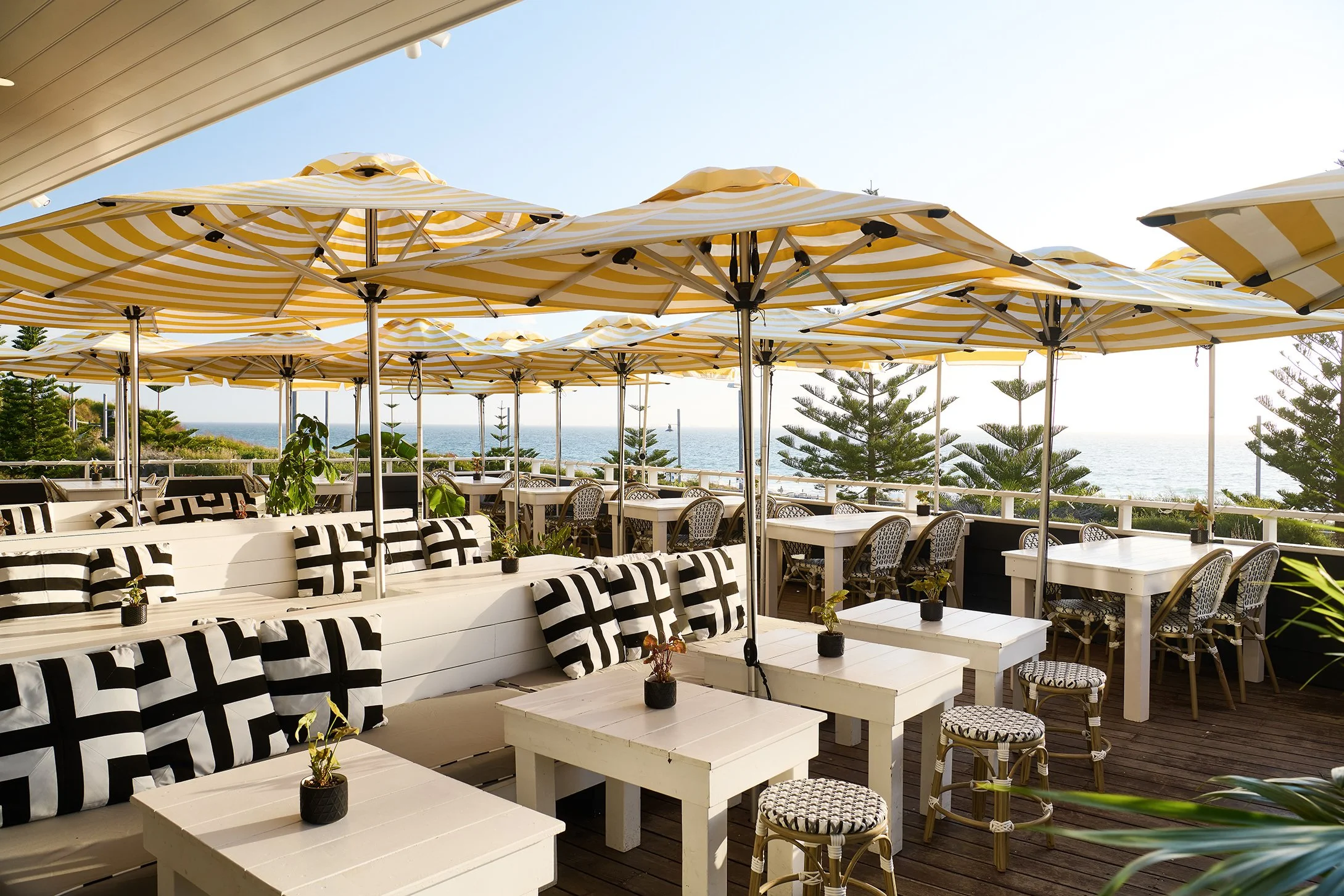 Outdoor dining area with white tables, black and white patterned pillows, and yellow and white striped umbrellas overlooking the ocean with pine trees in the background.