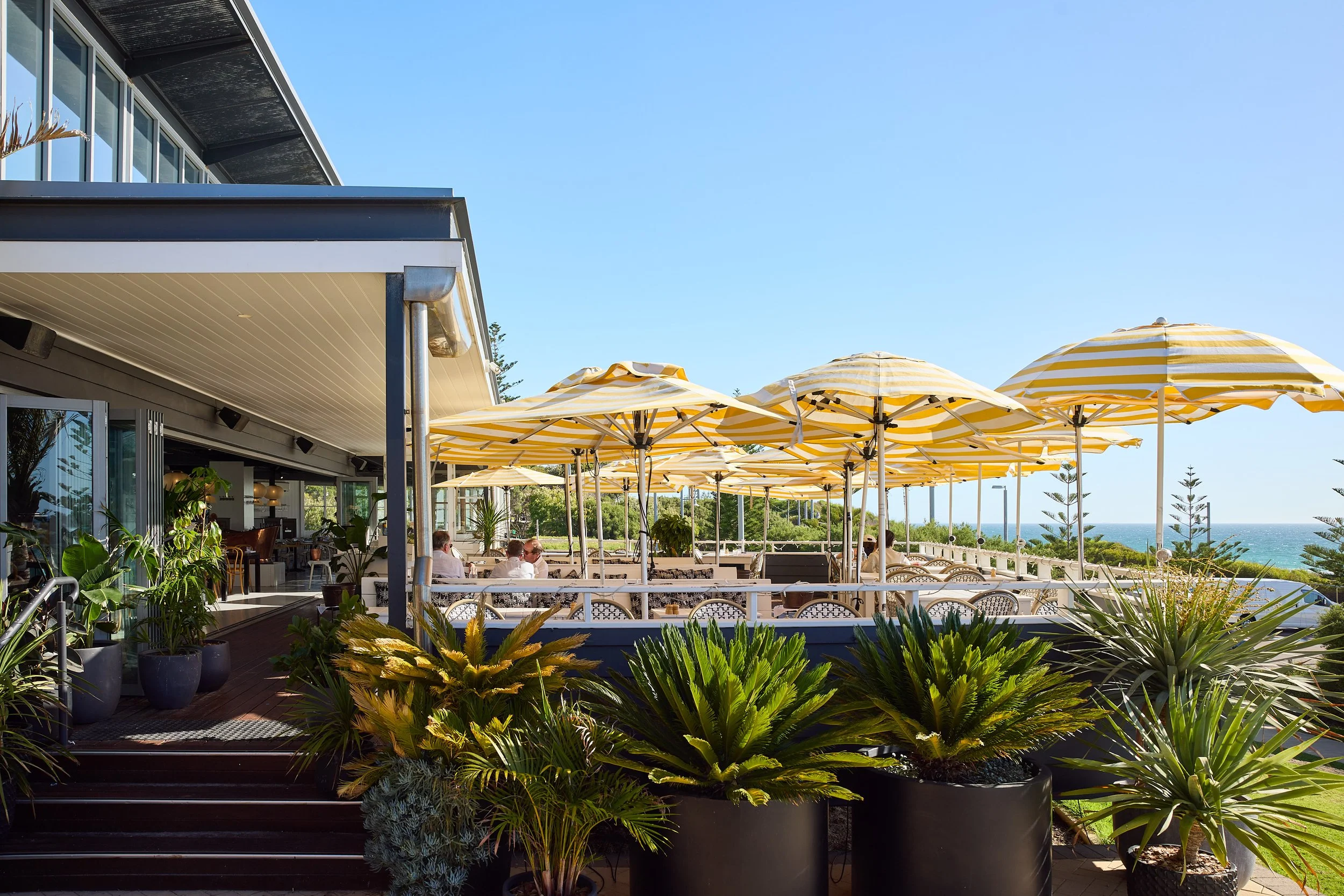 Outdoor patio with yellow and white striped umbrellas, green plants in pots, and people sitting at tables near the ocean under a clear blue sky.
