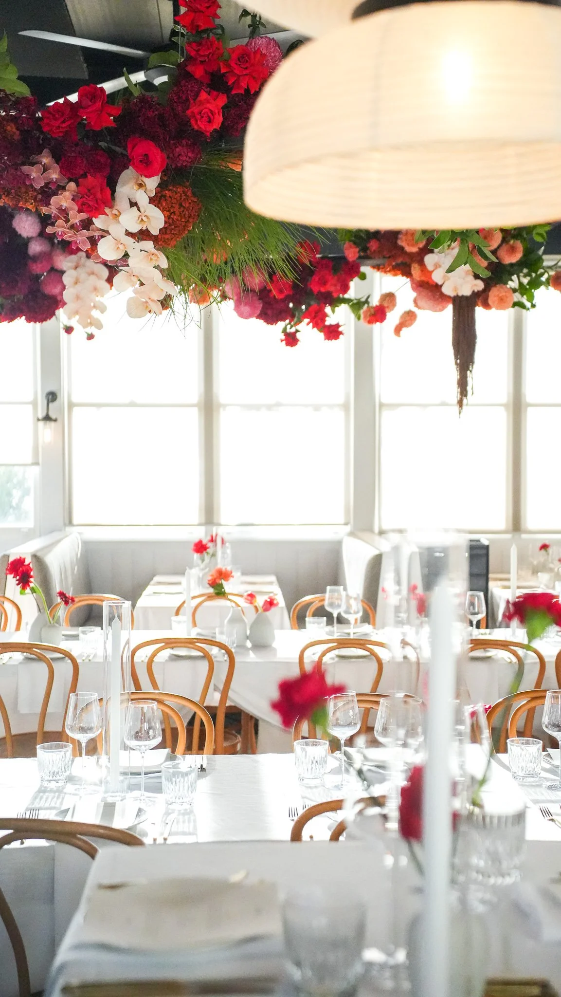 A beautifully decorated dining hall with tables covered in white tablecloths, elegant glassware, and floral centerpieces. The ceiling features a large floral arrangement of red, pink, and white flowers hanging above the tables, and natural light stre