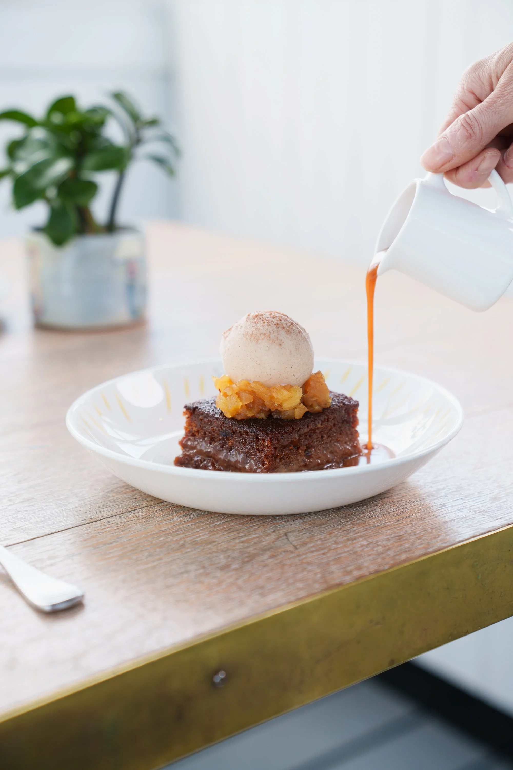 A slice of chocolate cake topped with a scoop of vanilla ice cream and caramel sauce being poured over it on a white plate. There is a potted plant with green leaves in the background.