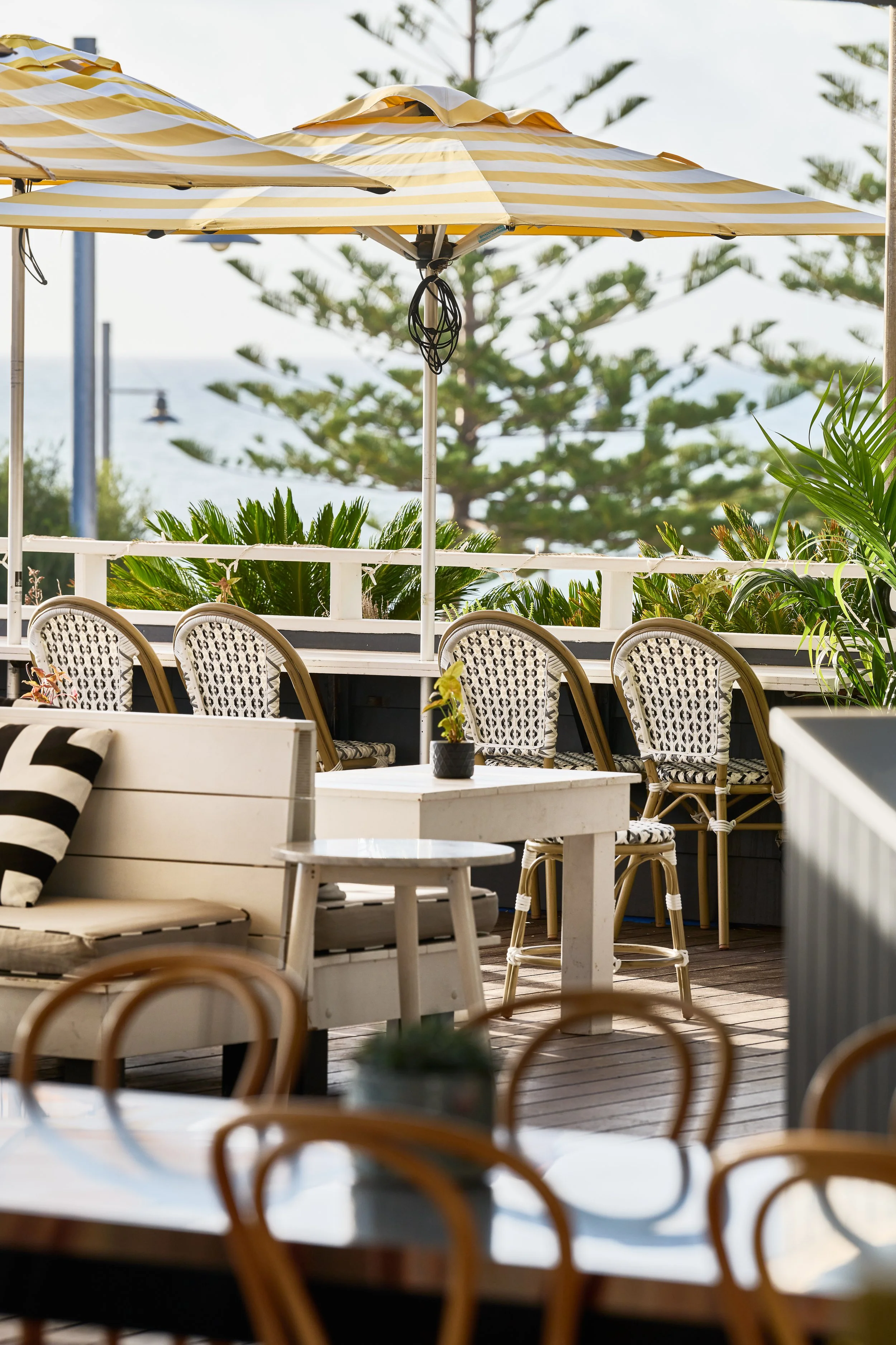 Outdoor patio with yellow and white striped umbrellas, white chairs with black and white patterned cushions, and greenery in the background.