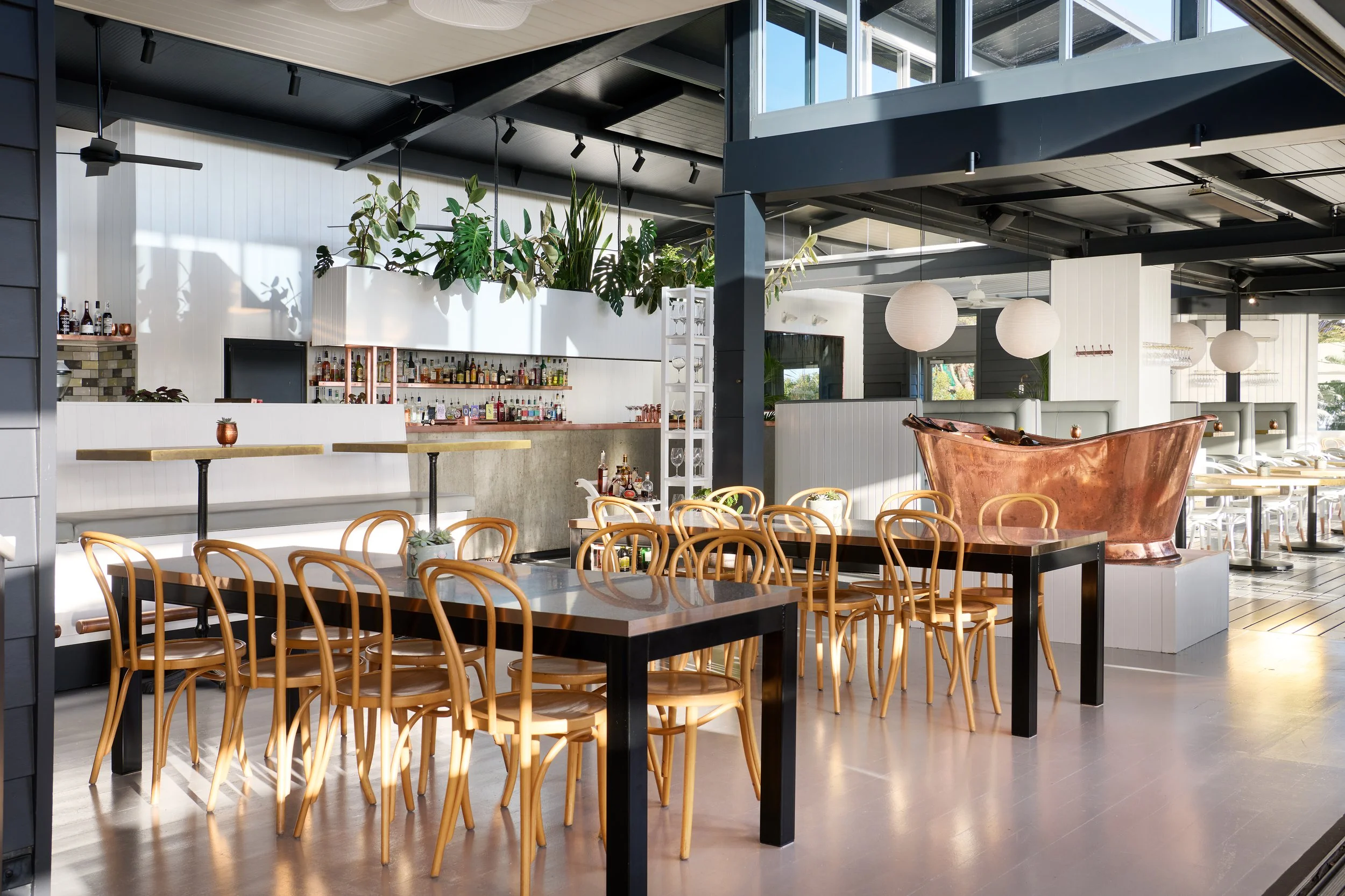 Inside view of a modern restaurant with wooden chairs, black tables, a bar area with bottles, and hanging round paper lantern lights, with plants and large windows letting in natural light.