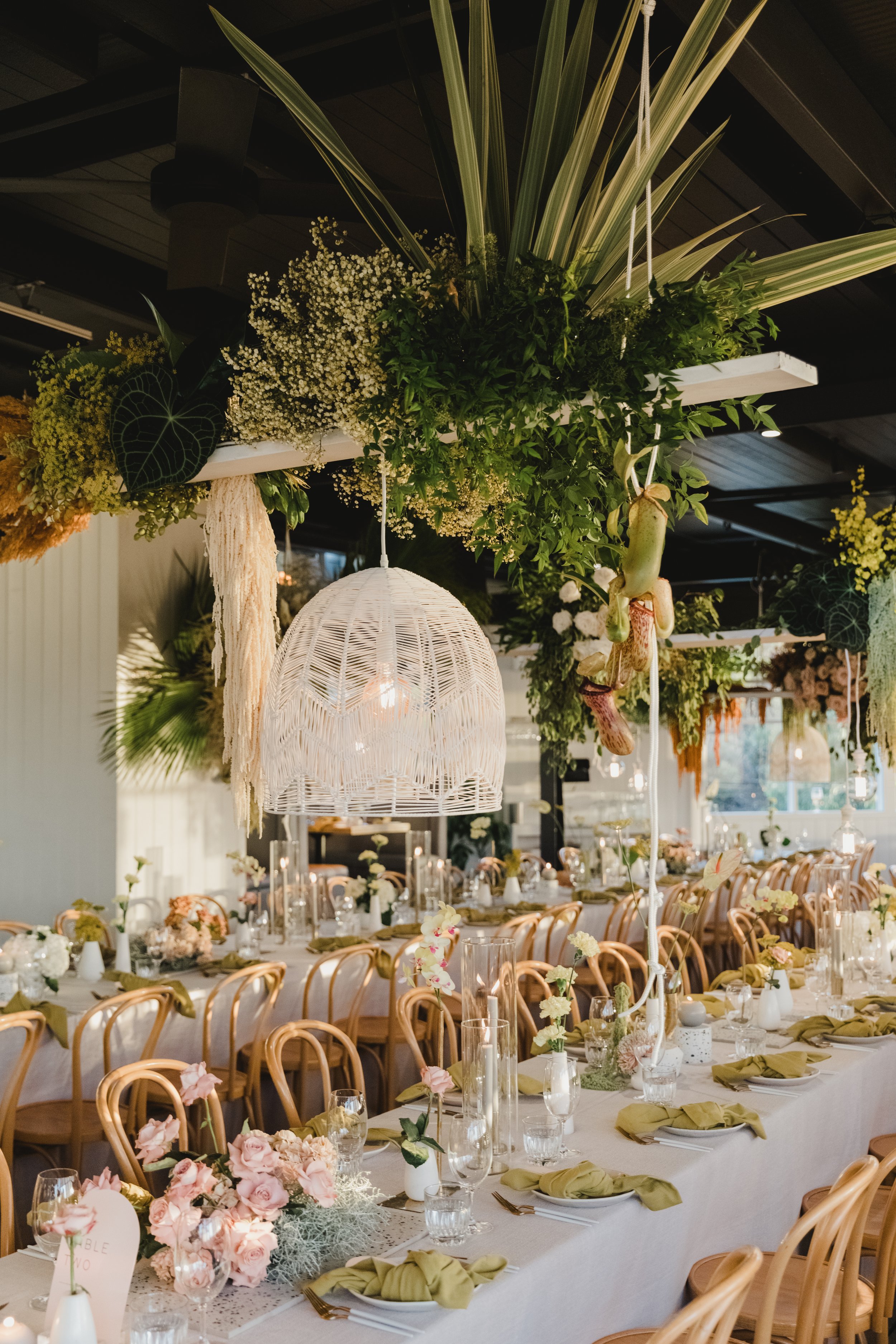 Elegant indoor wedding reception decorated with floral arrangements and greenery, featuring long banquet tables, light wood chairs, and hanging white woven light fixtures.